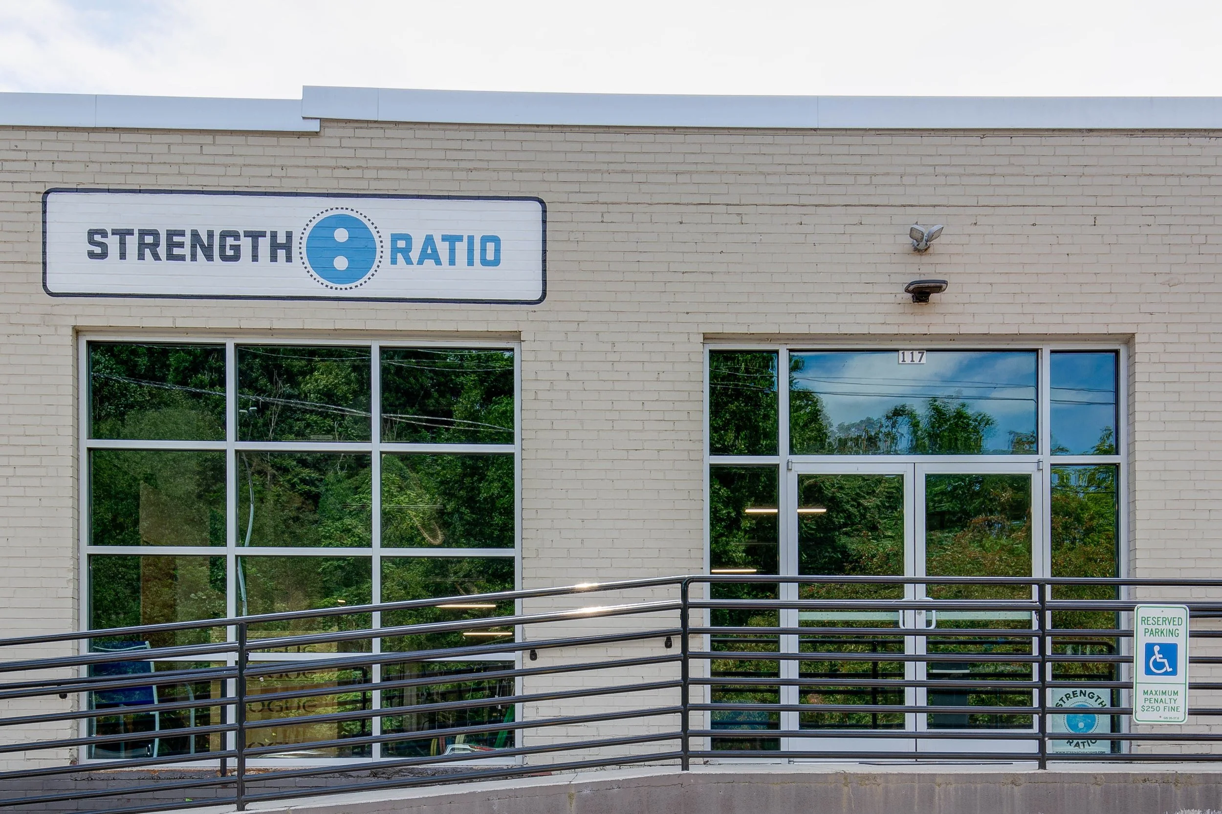 Front of a building with a sign reading 'Strength 8 Ratio' and large windows reflecting trees. There's a wheelchair parking sign and a security camera on the wall.