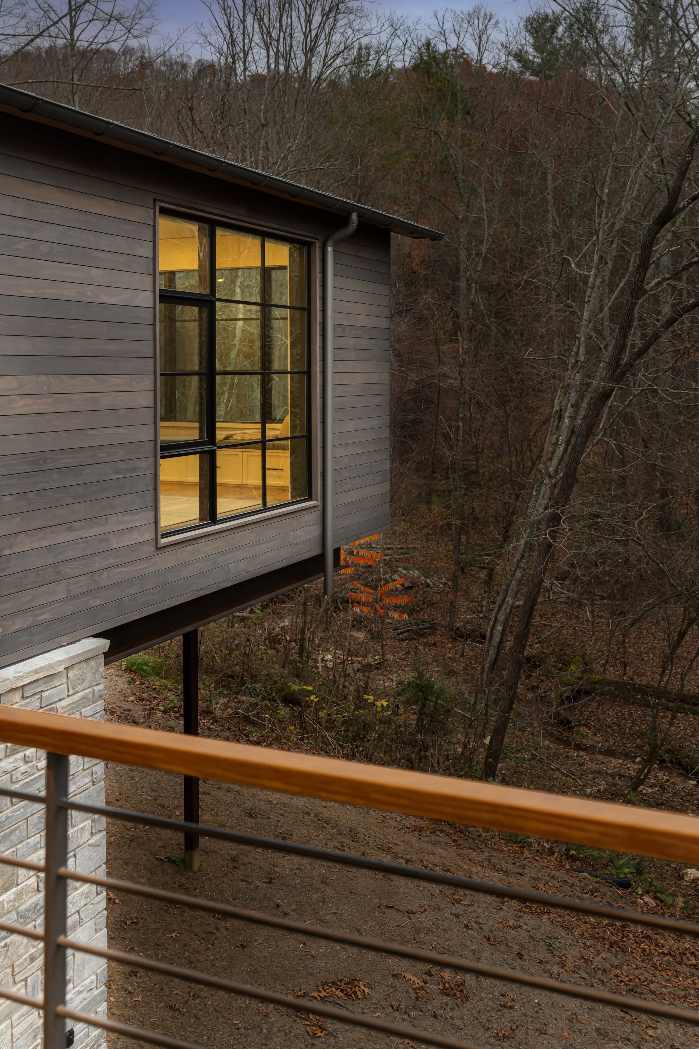 View of a modern house overlooking a wooded area, with a large window showing a cozy interior. A wooden railing is in the foreground, and leafless trees are visible in the background.