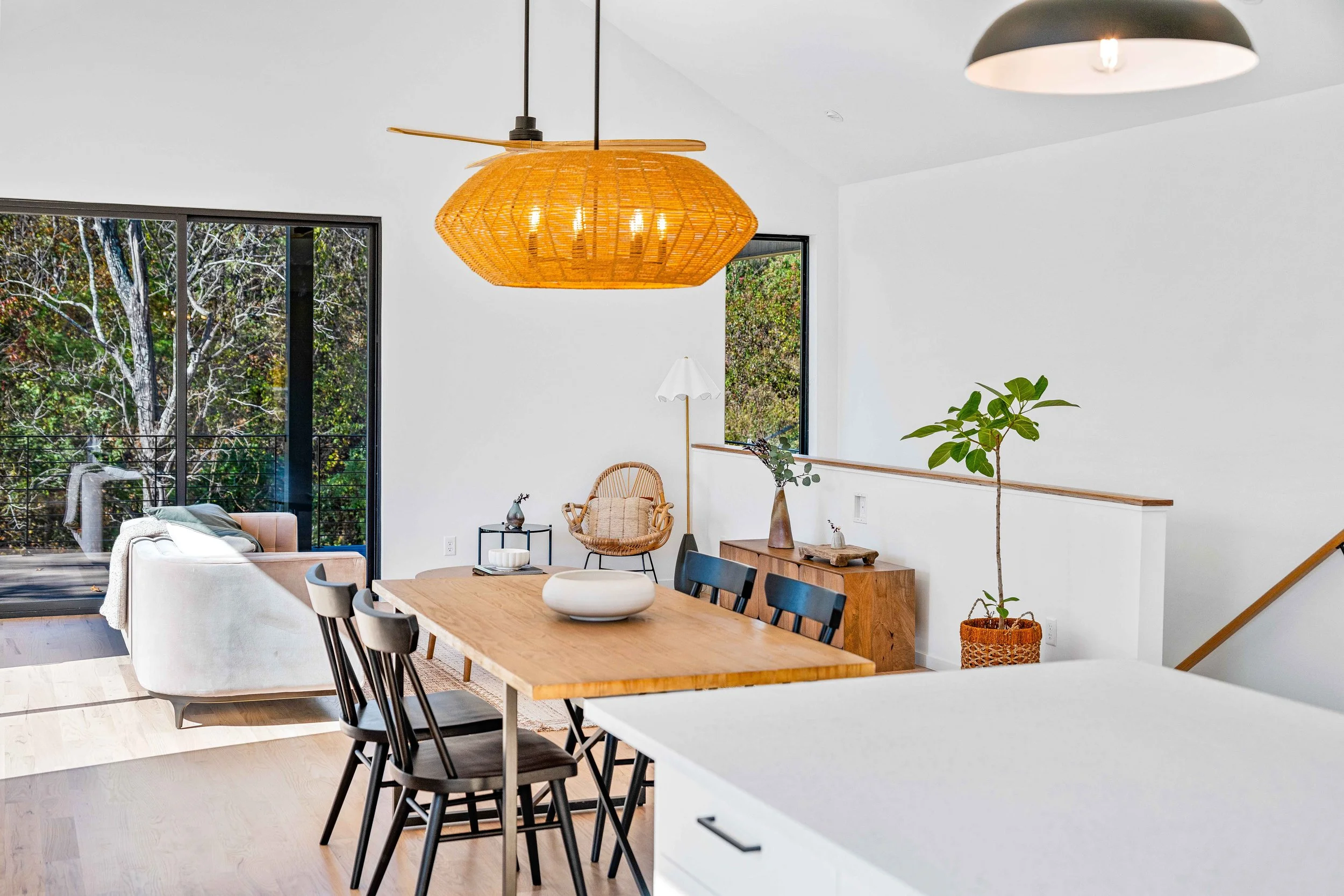 A modern open-concept living and dining area with large windows, a dining table with black chairs, a white sofa, a wooden sideboard with decorative items, potted plants, and a large woven pendant light.