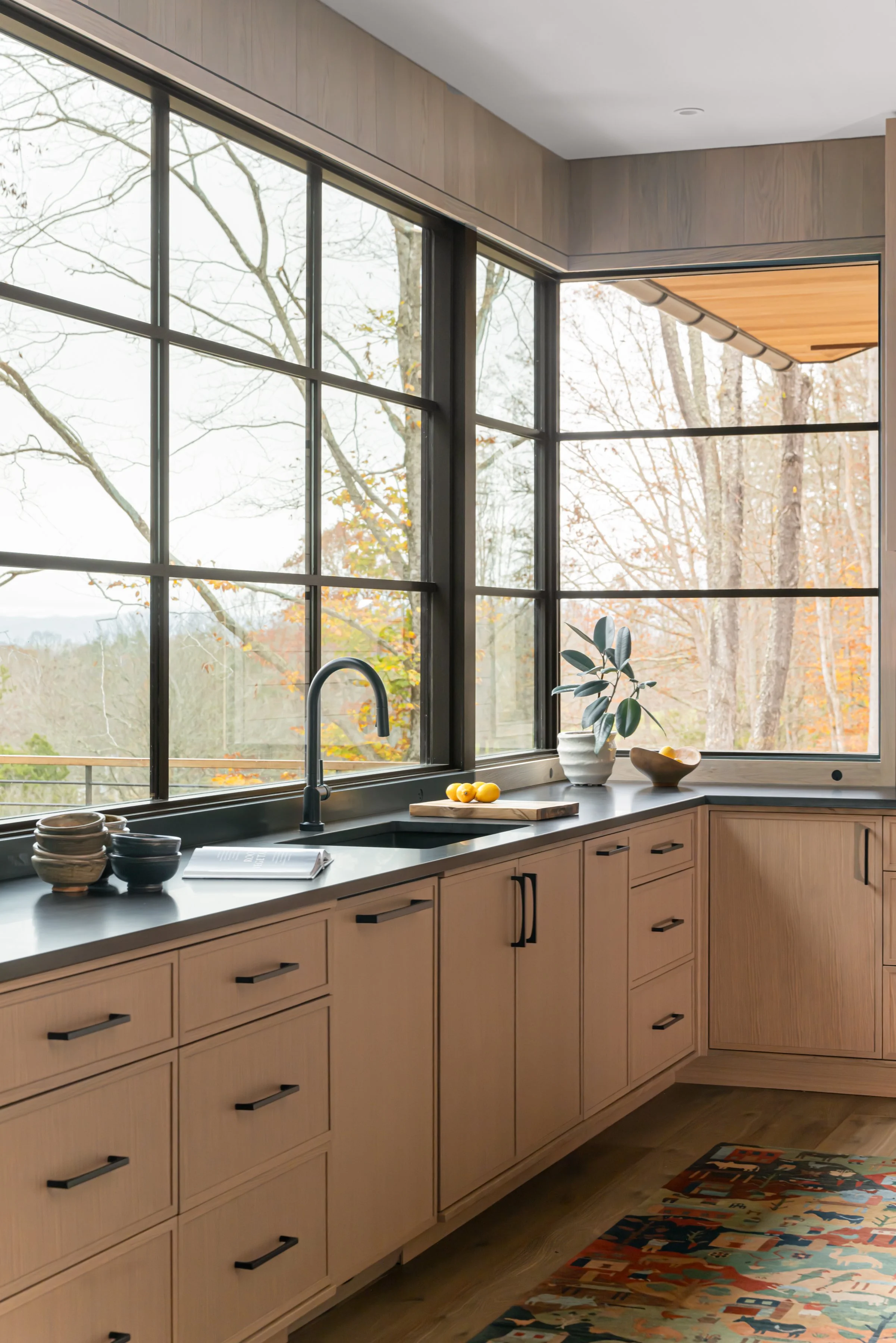 Modern kitchen with large windows overlooking a wooded outdoor scene, light wood cabinetry, a black sink faucet, bowls and a cutting board on the countertop, a potted plant, and a colorful patterned rug.