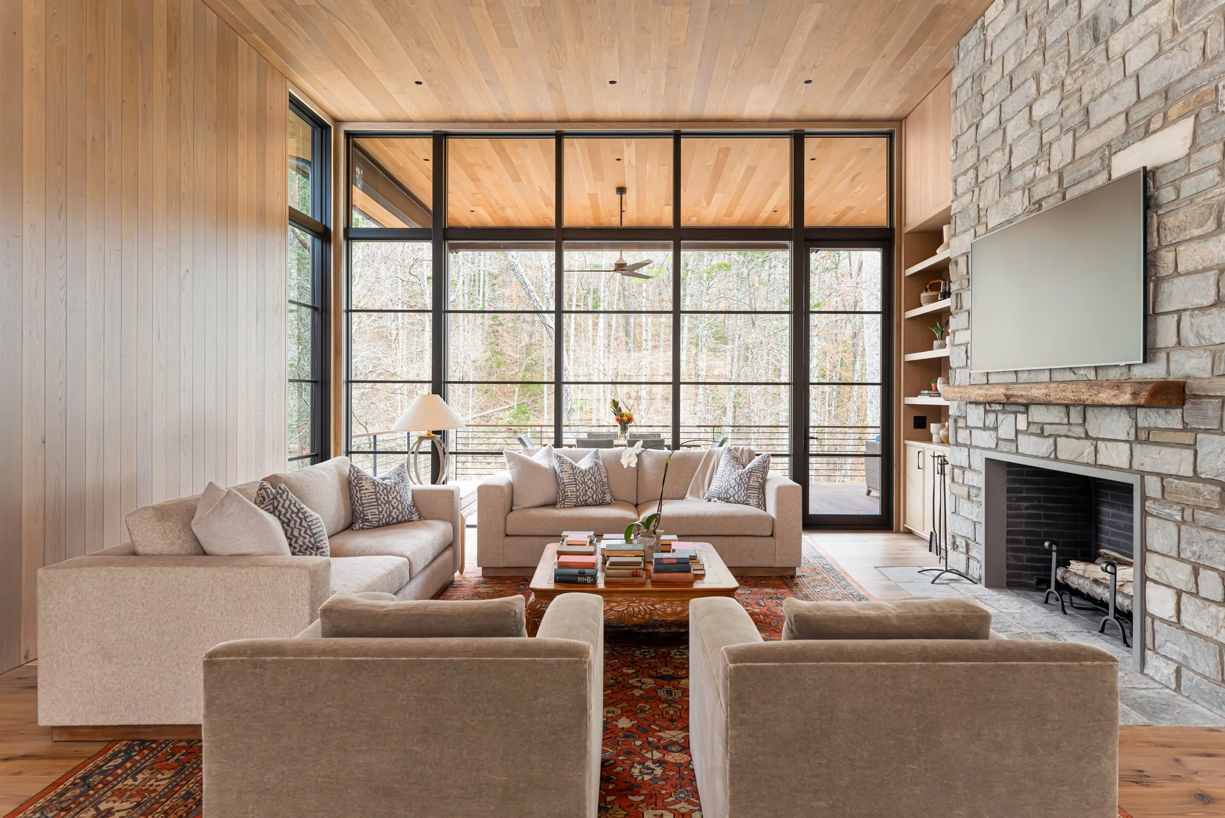 A modern living room with beige sofas, a wooden coffee table with books and a vase with flowers, a rug, a stone fireplace, and large windows showing a forest outside.