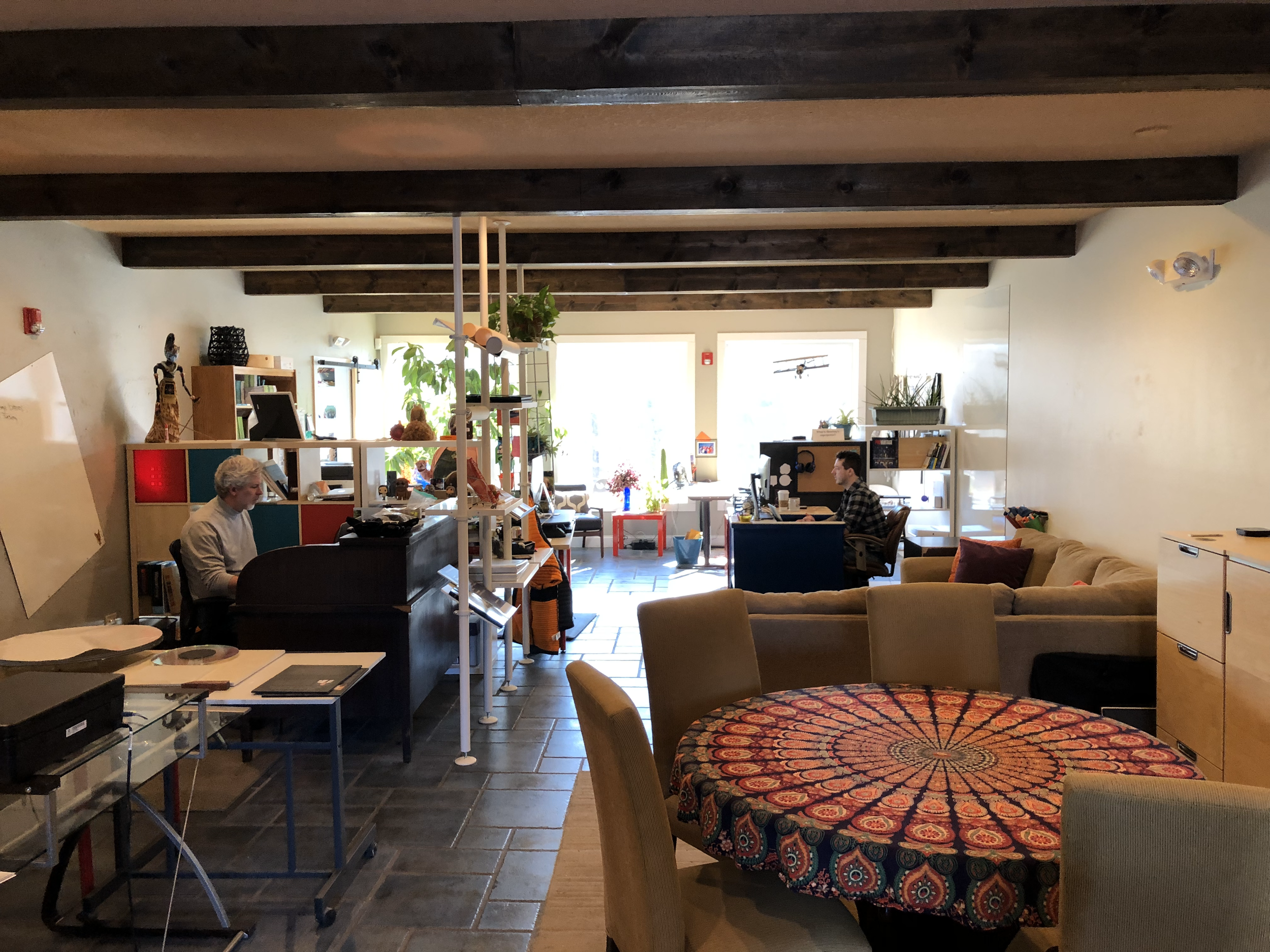 An indoor workspace with a round table covered by a colorful, patterned cloth in the foreground, beige chairs, a sofa with pillows, a person working on a computer at a desk, a woman playing the piano, and various books and decorations, with natural l