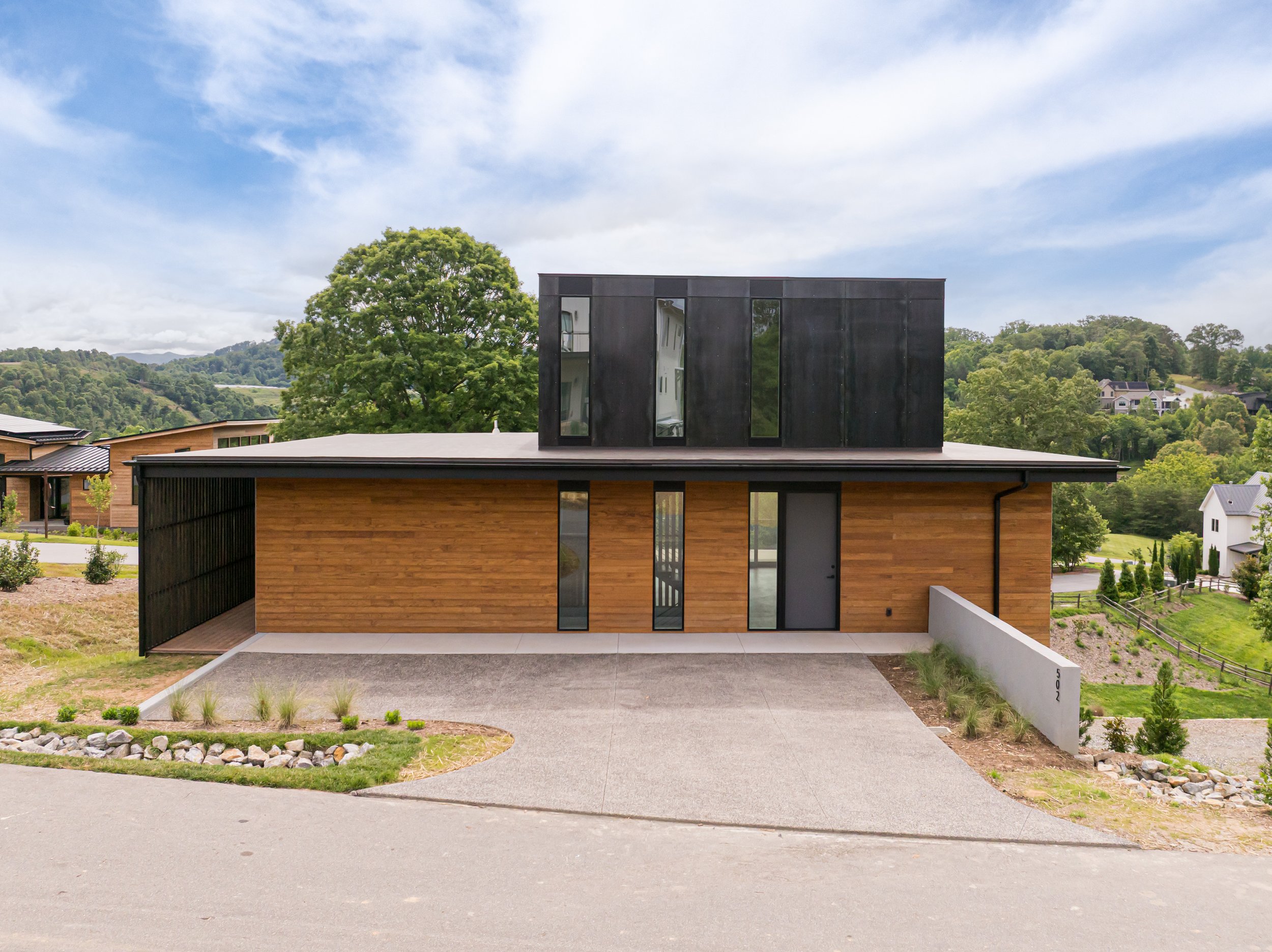 Modern house with wood and black exterior, driveway, and surrounding greenery, located in a hilly area with trees and other houses in the background.