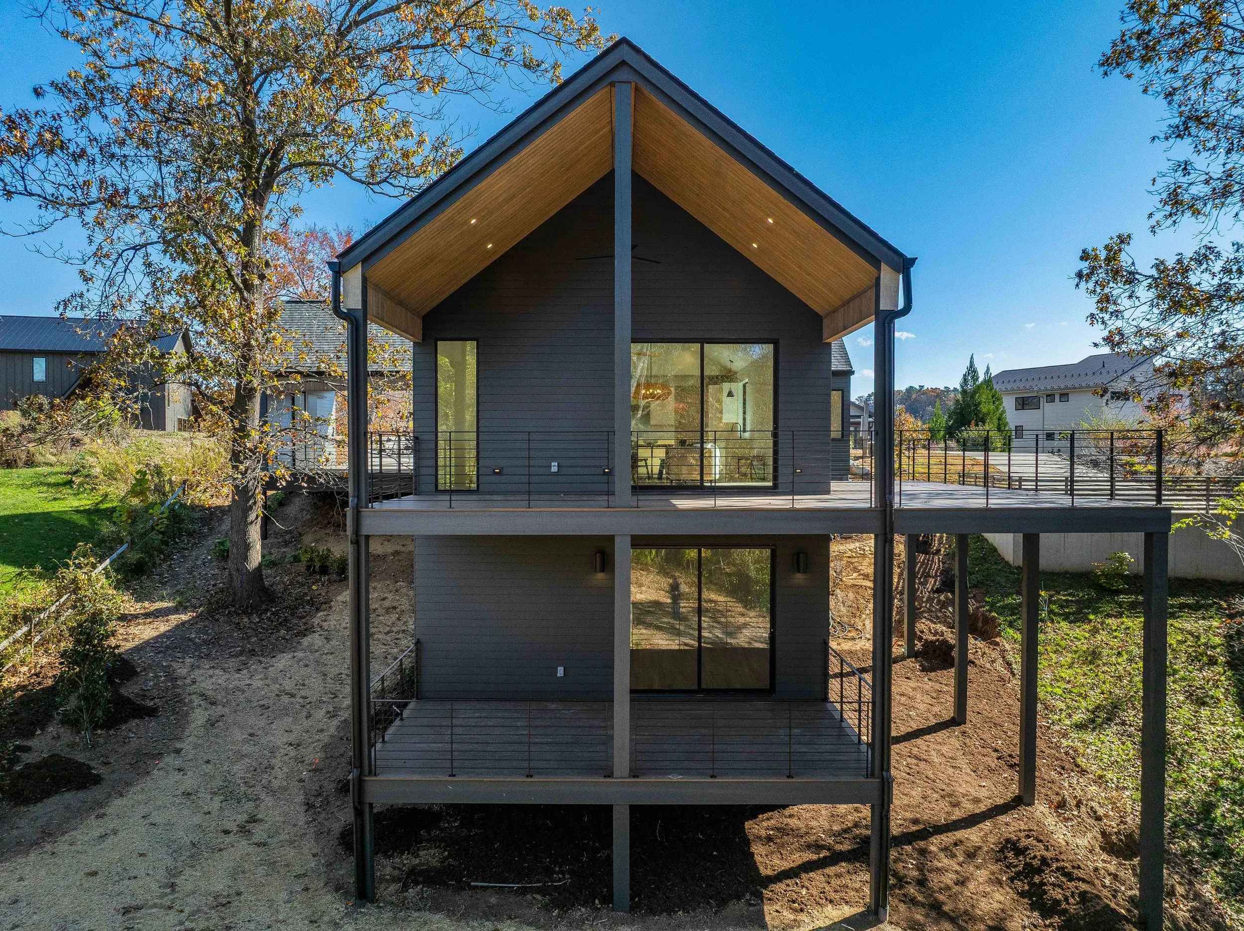 Modern two-story house with large glass doors and dark siding, surrounded by trees and a landscape with partly cloudy sky.