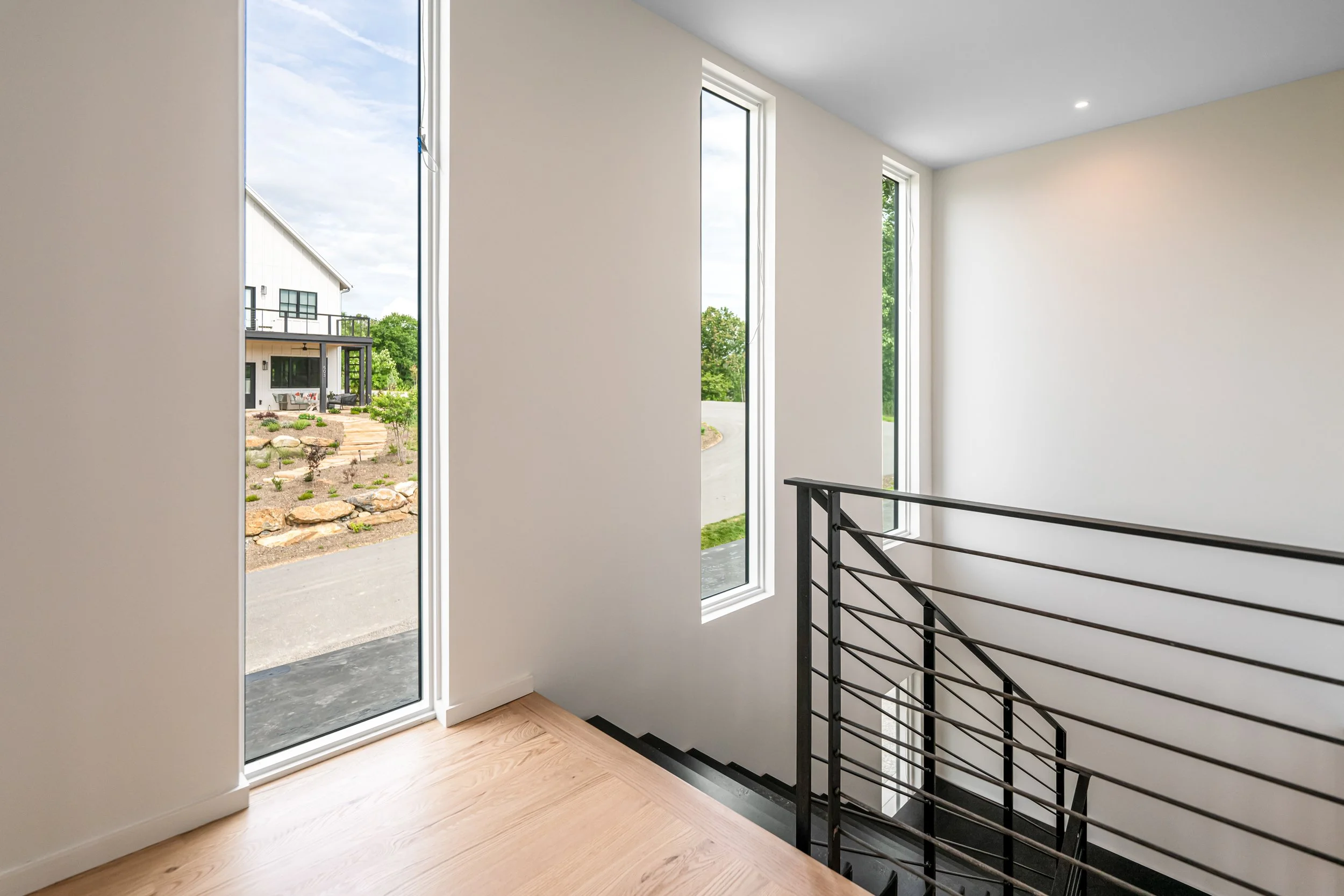 Interior view of a modern house with large windows, a staircase with metal railing, and a view of a landscaped garden outside.