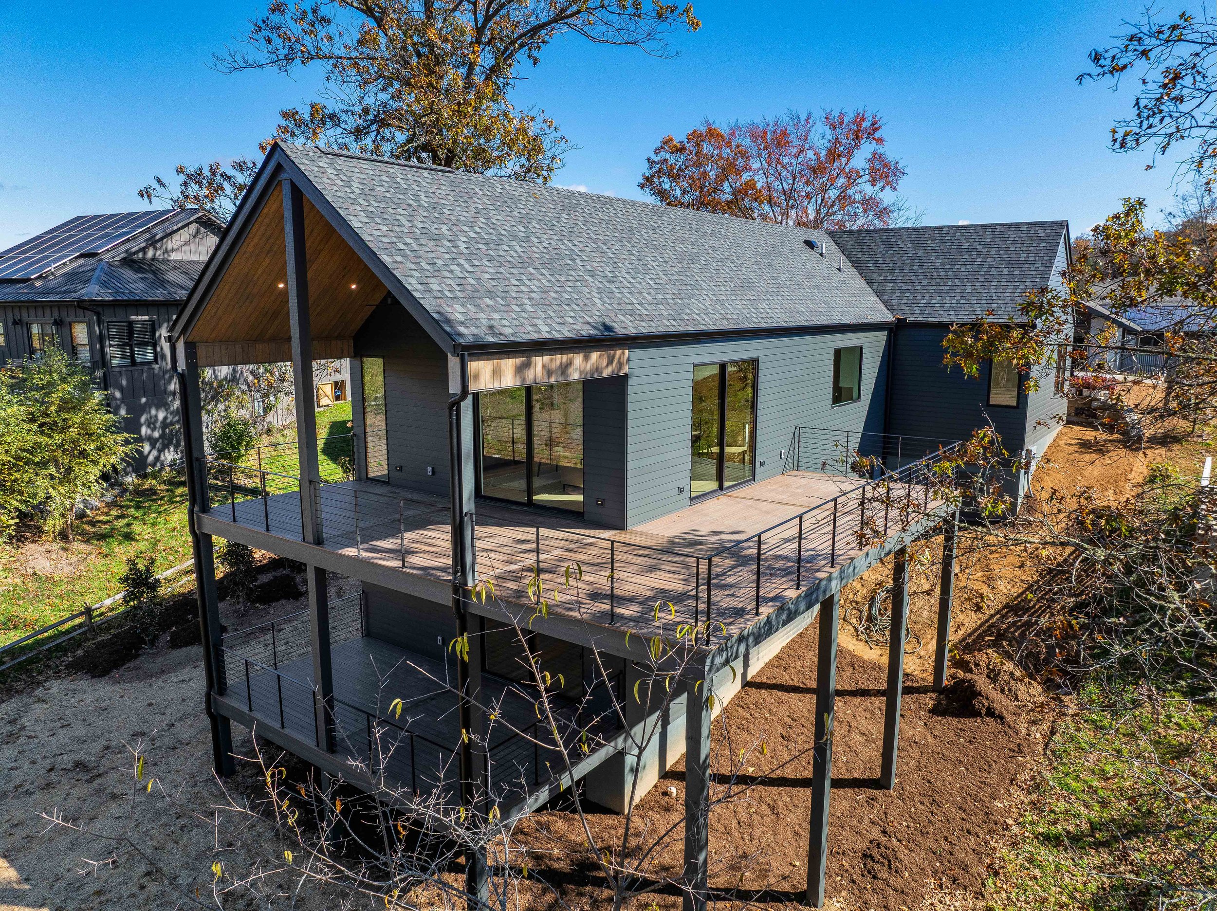 A modern house with a large wooden deck, multiple sliding glass doors, and gray siding, situated on stilts with trees and neighboring houses nearby.