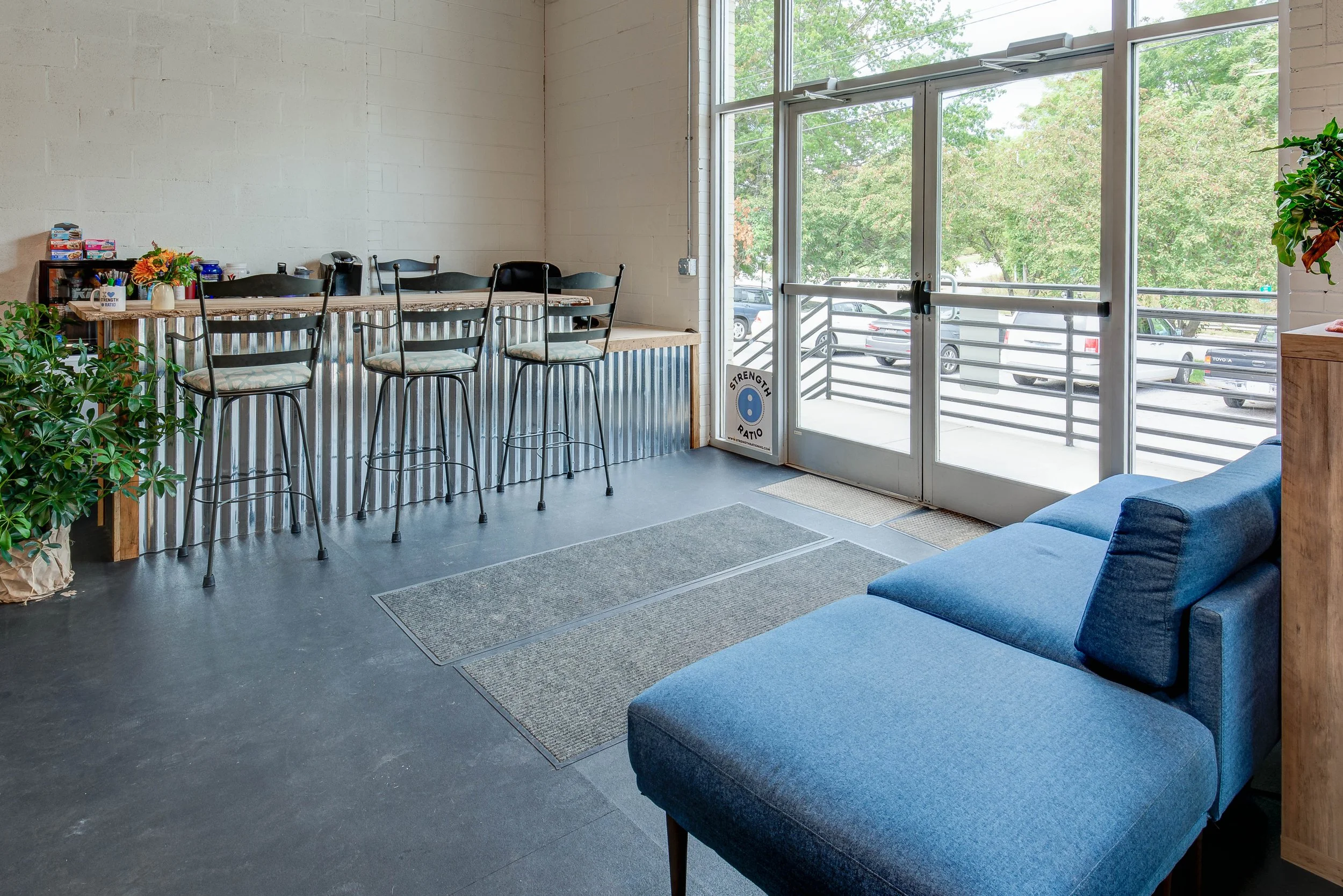 Interior of a modern room with a blue couch near glass doors leading outside, a high wooden counter with metal panels, behind which are four black barstools, and a small shelf with snacks and flowers, greenery on the side, and parking lot visible thr