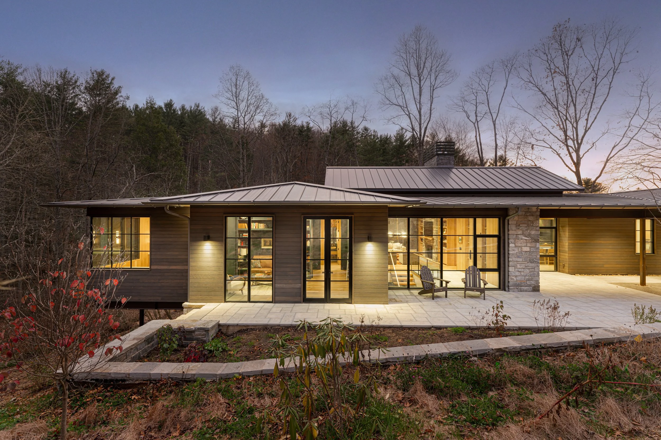 A modern house with large glass doors and windows, featuring outdoor seating on a stone patio, surrounded by leafless trees and forest, during dusk.