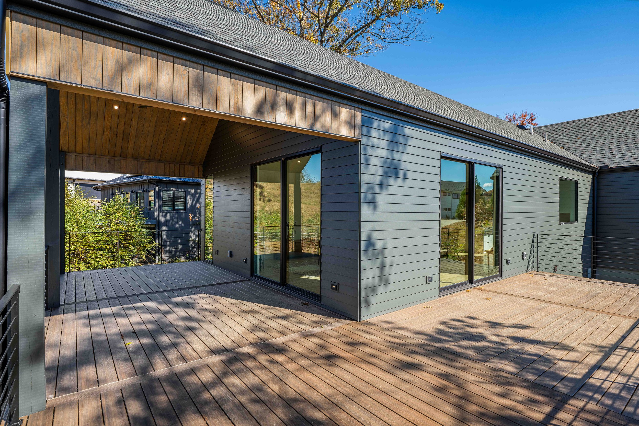 Modern house balcony with wooden floor, glass sliding doors, and partial roof overhead, with trees and other houses in the background under a clear blue sky.