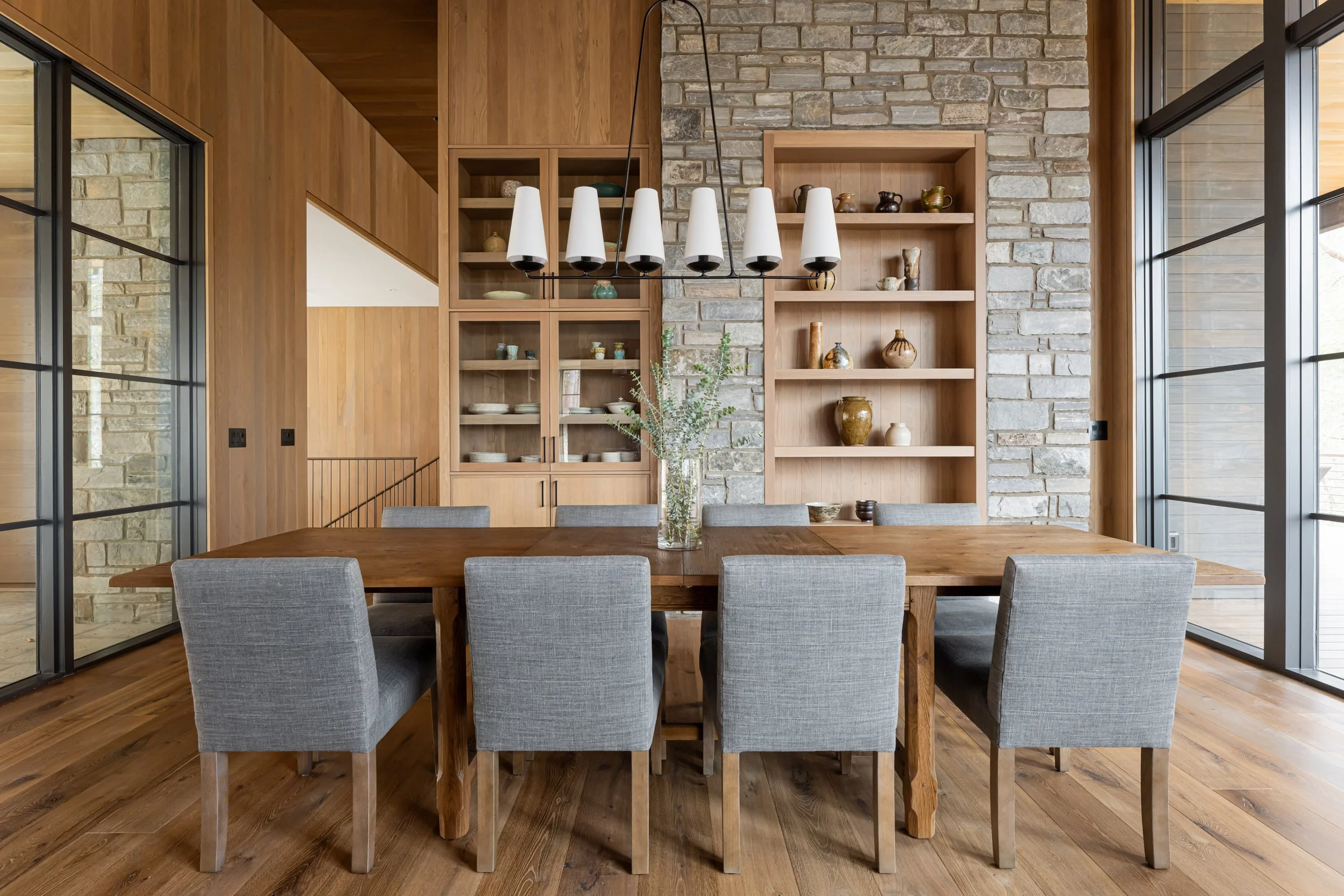 A modern dining room with a large wooden table, eight gray upholstered chairs, a glass vase with green foliage, wooden shelves with decorative pottery, and large windows with black frames, featuring a stone and wood interior design.
