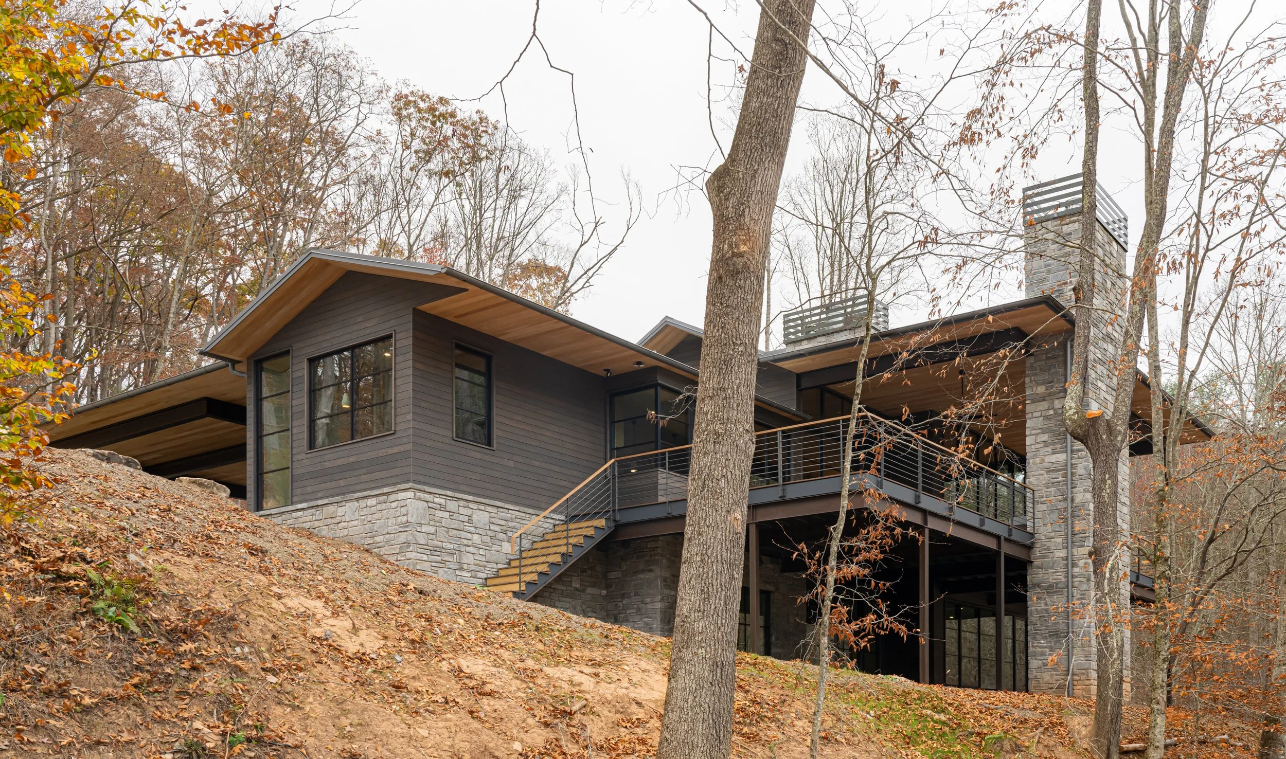 Modern house on a hillside in a forest with fall foliage, featuring dark wooden siding, large windows, stone accents, and a balcony with metal railing.