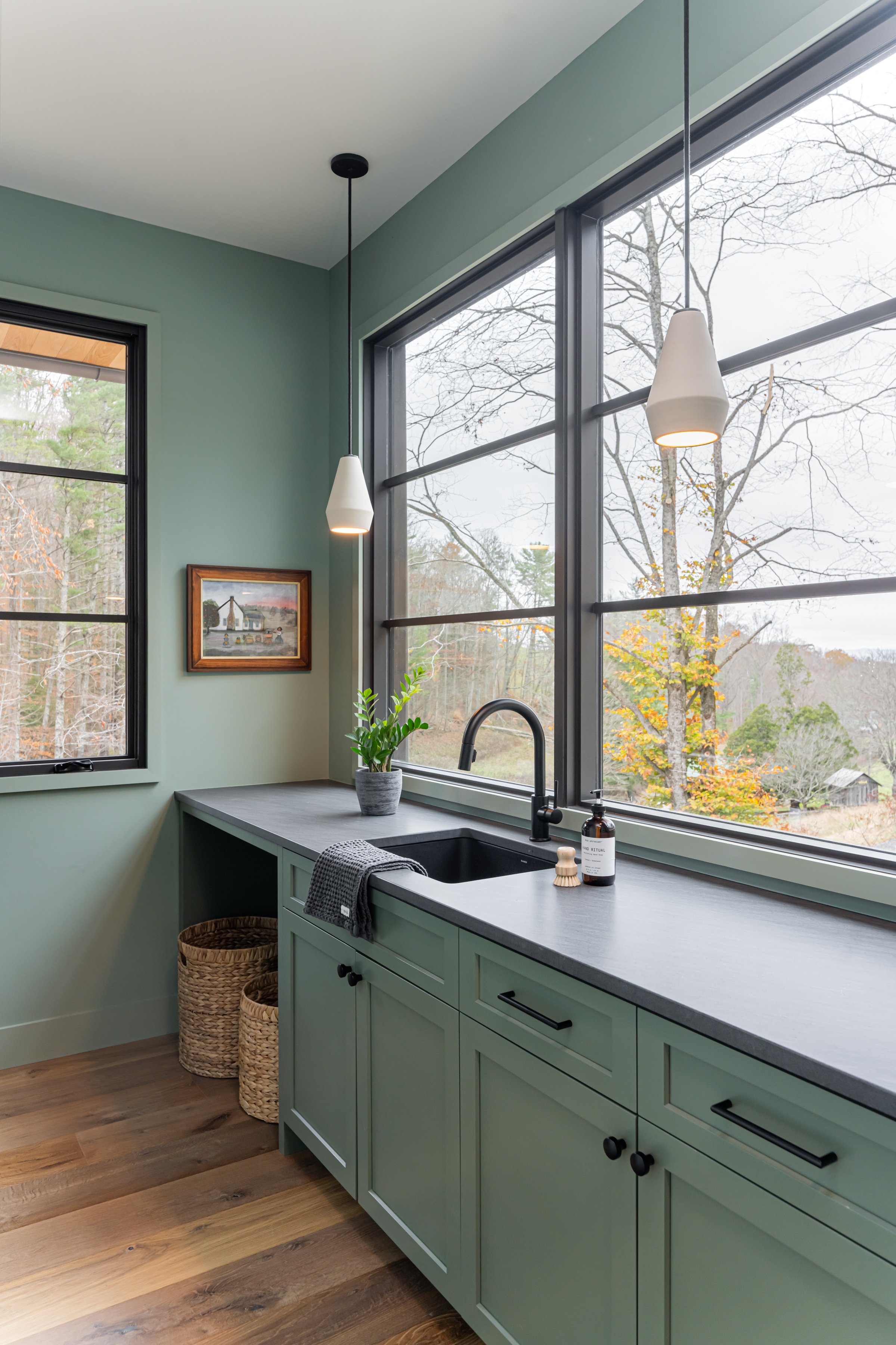 Bright kitchen with large windows, mint green cabinetry, black hardware and fixtures, wooden floors, a small plant, and a black sink, with outdoor trees visible through the windows.
