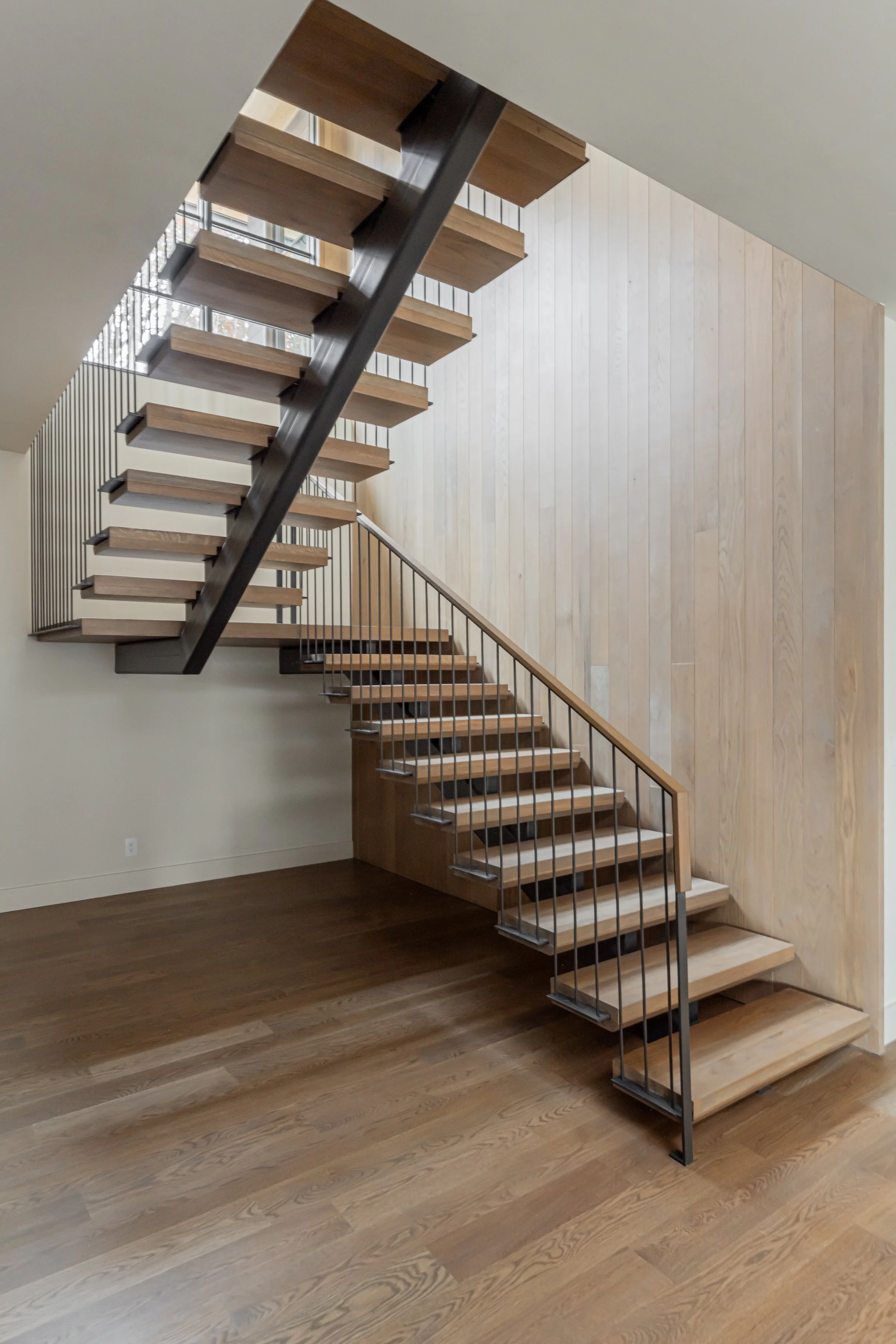 Interior view of a modern staircase with wooden steps, metal railing, and a wooden wall, leading upstairs in a house or building.