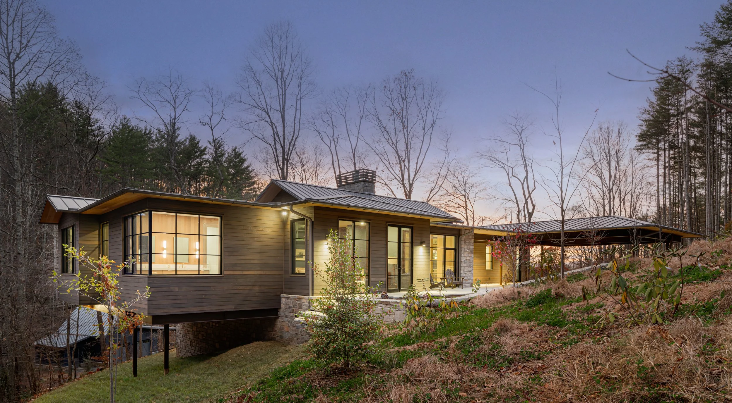 A modern house with large windows, situated on a hillside surrounded by trees, during sunset.