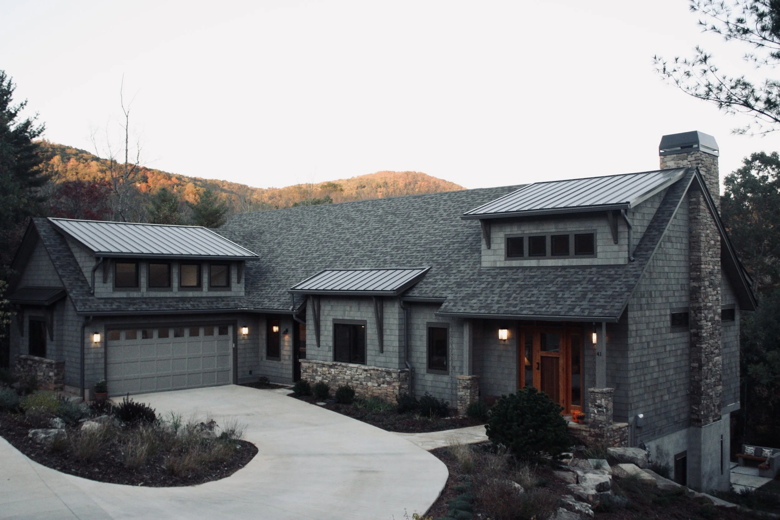 A modern house with gray shingle siding, stone accents, and a metal roof, situated on a sloped landscape with trees and mountains in the background.