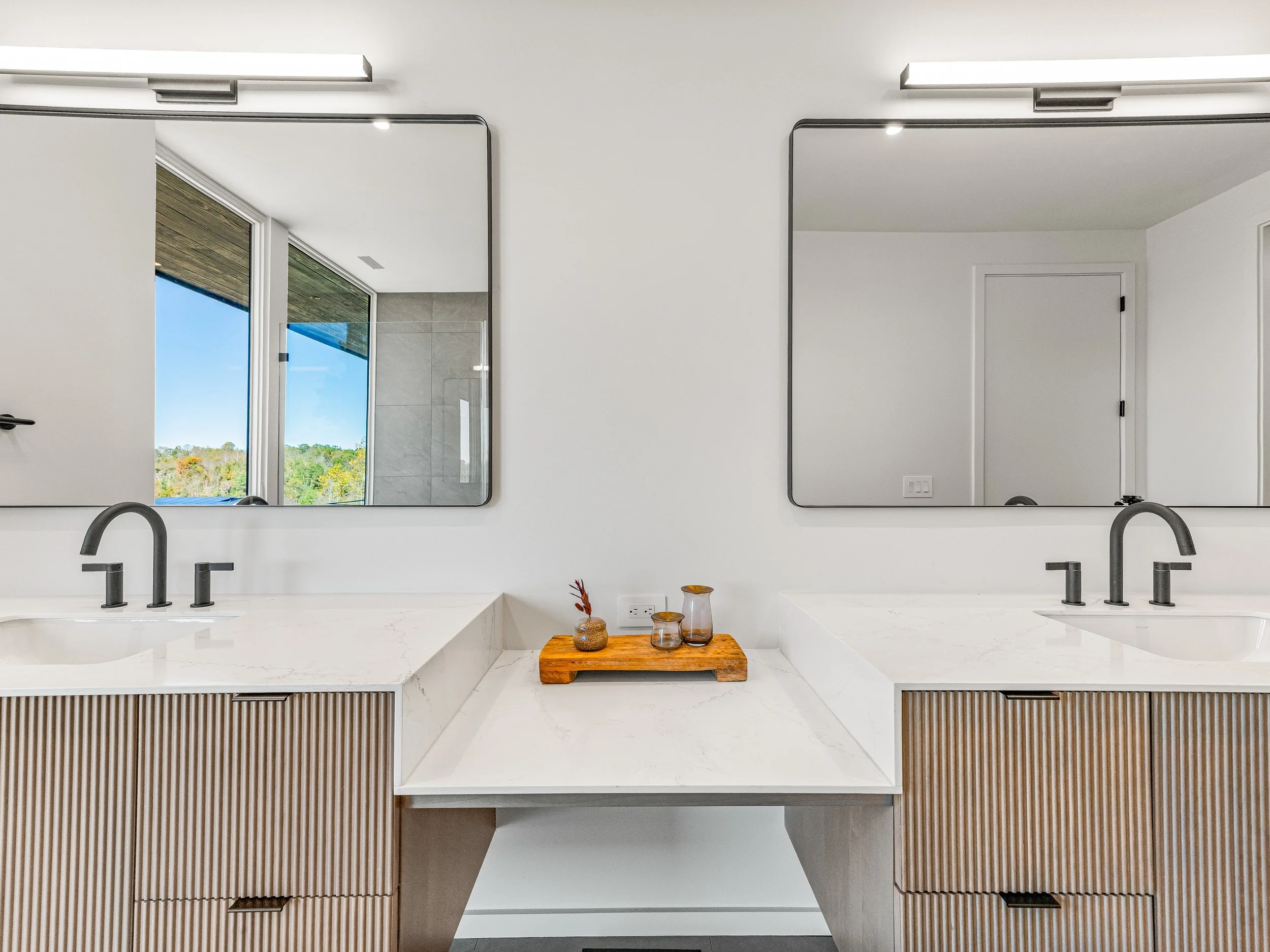 Modern bathroom double vanity with large mirrors, white countertops, black faucets, and a small wooden tray with decorative items, bright natural light from windows.
