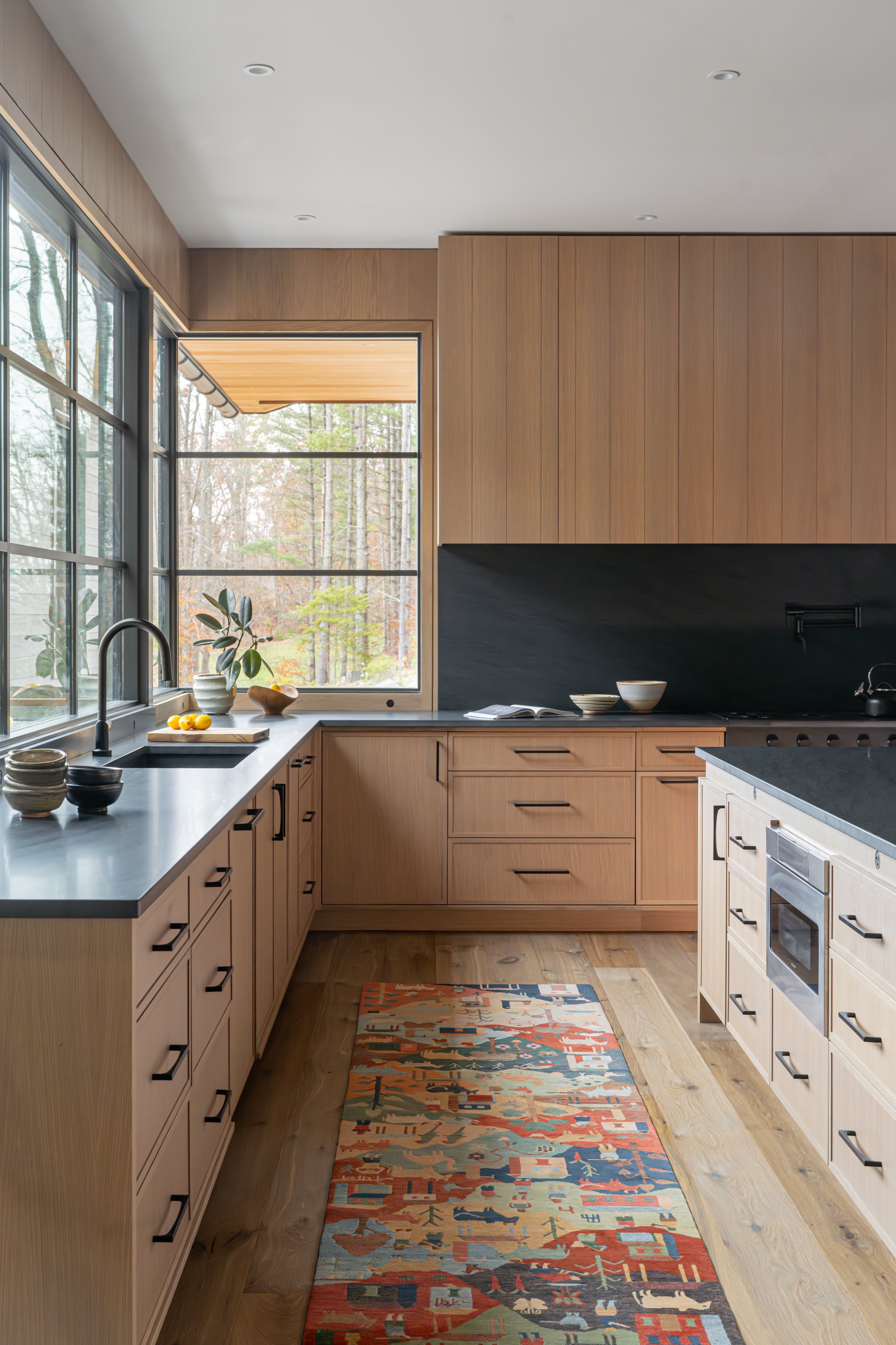Modern kitchen with wooden cabinets, black countertops, a window with a view of trees, and decorative bowls and a plant on the counter.