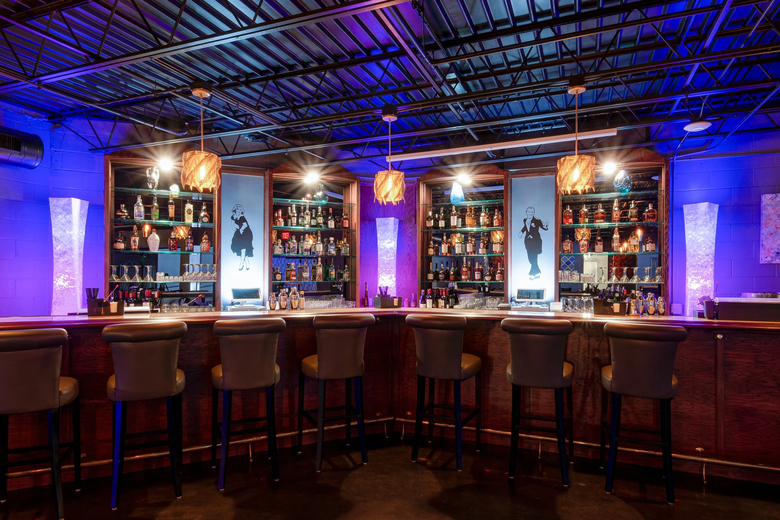 A bar with a curved wooden counter, seven chairs, purple and blue lighting, hanging pendant lights, shelves with various liquor bottles, and black and white silhouette images of a woman and a man in the background.