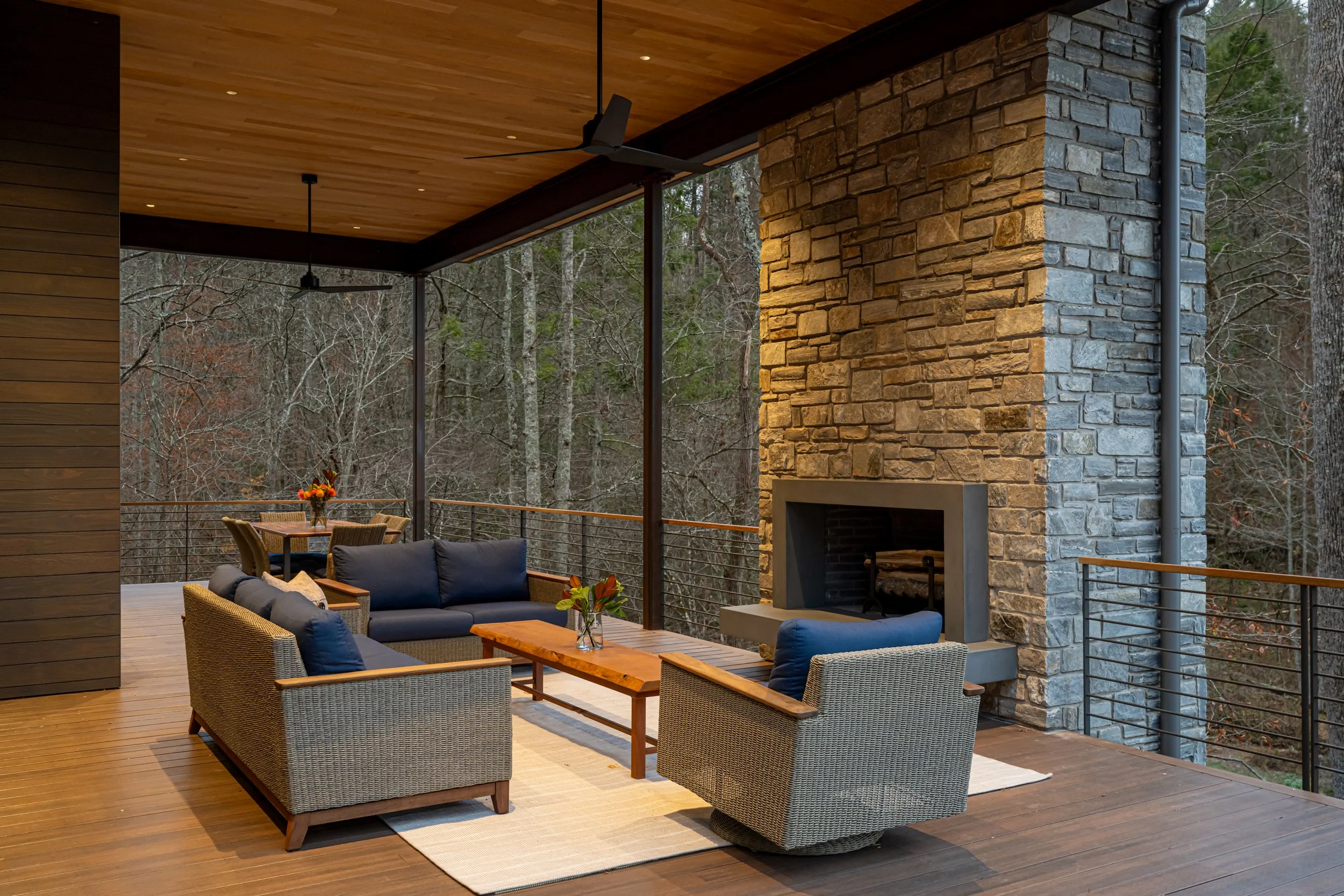 Covered outdoor living space with wooden ceiling, stone fireplace, wicker and wooden furniture, and a view of trees in the woods.