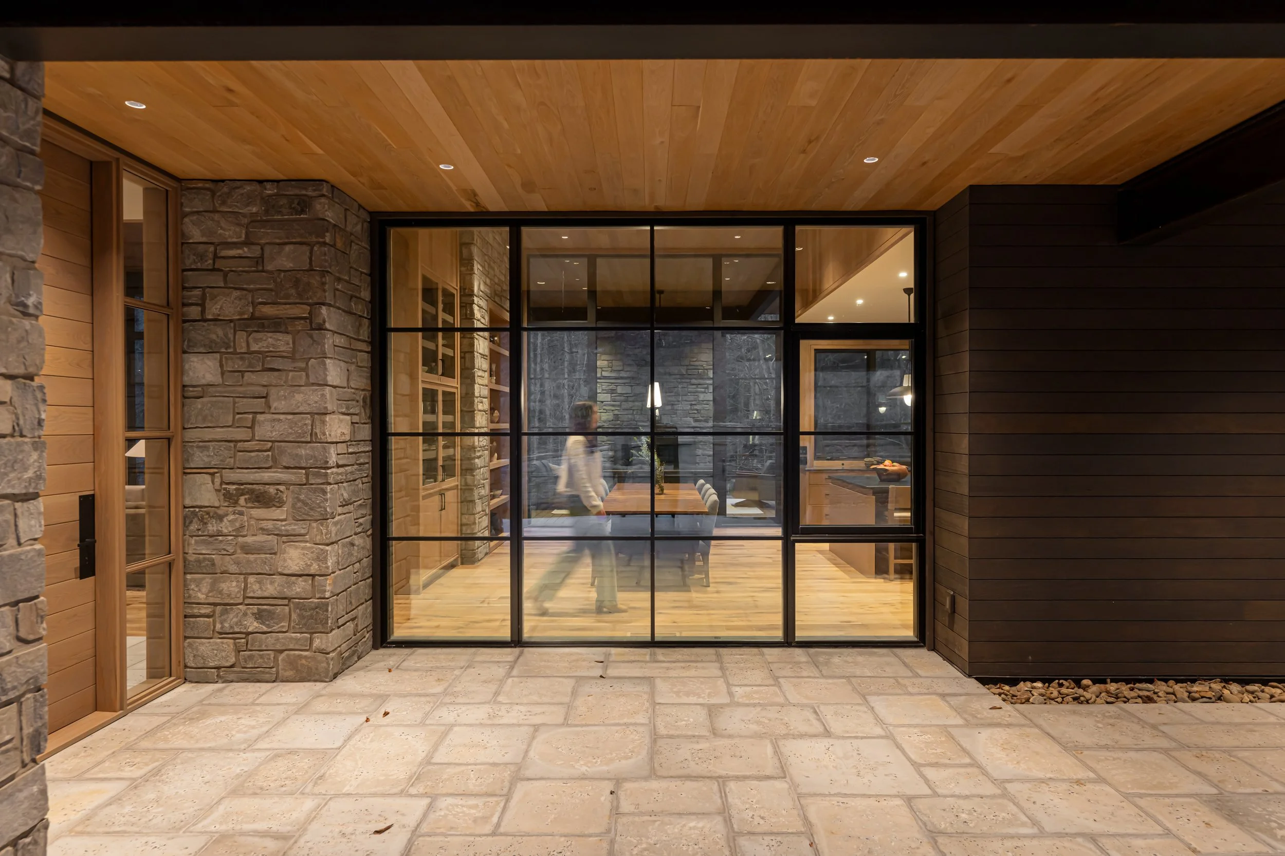 Interior view through large glass door showing a dining room with a wooden table, chairs, and stone and wood interior walls, and a woman walking inside.