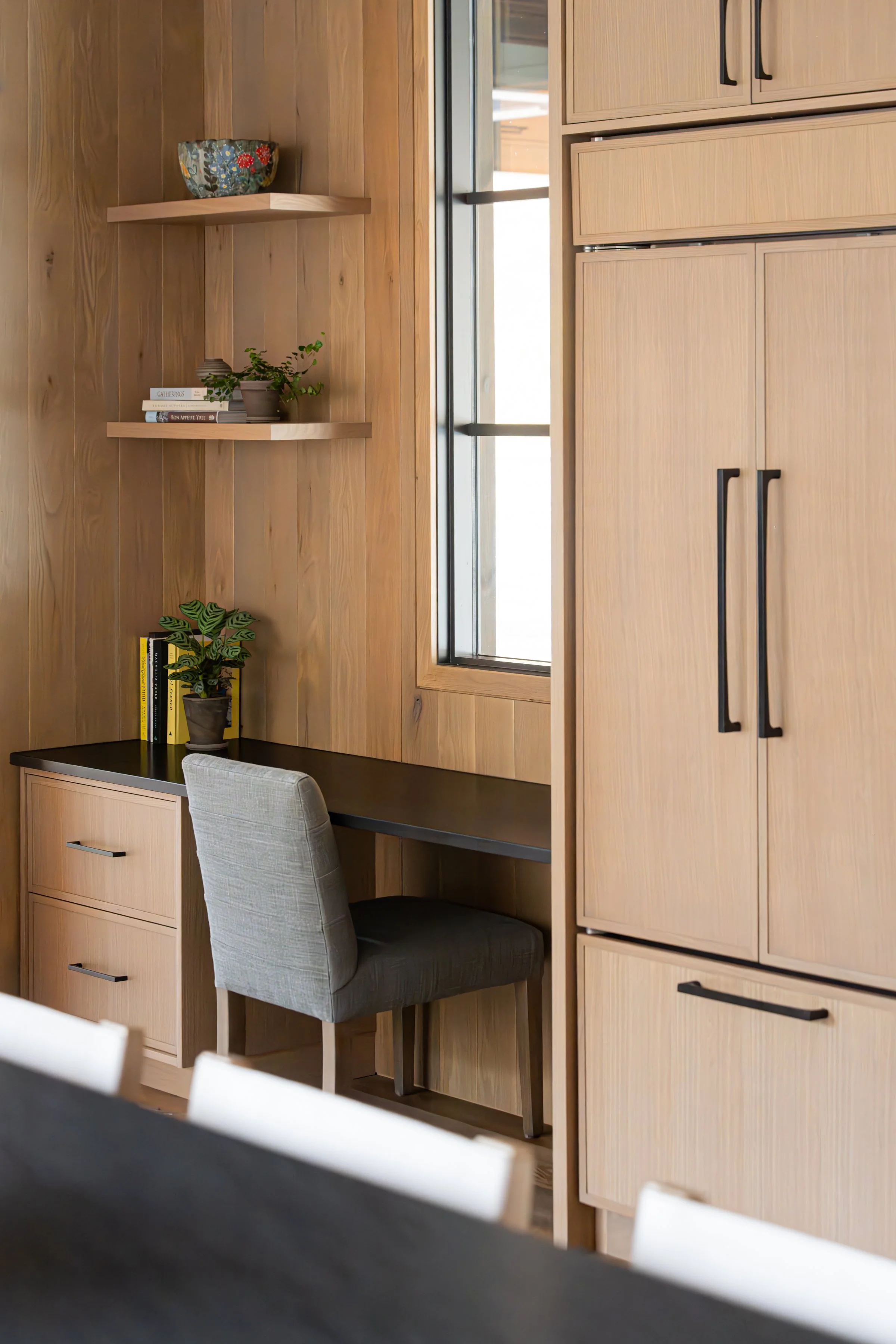 A small workspace with a built-in desk, grey upholstered chair, wooden shelves with books and decorative items, a potted plant, and a large window, all in a wood-paneled room.