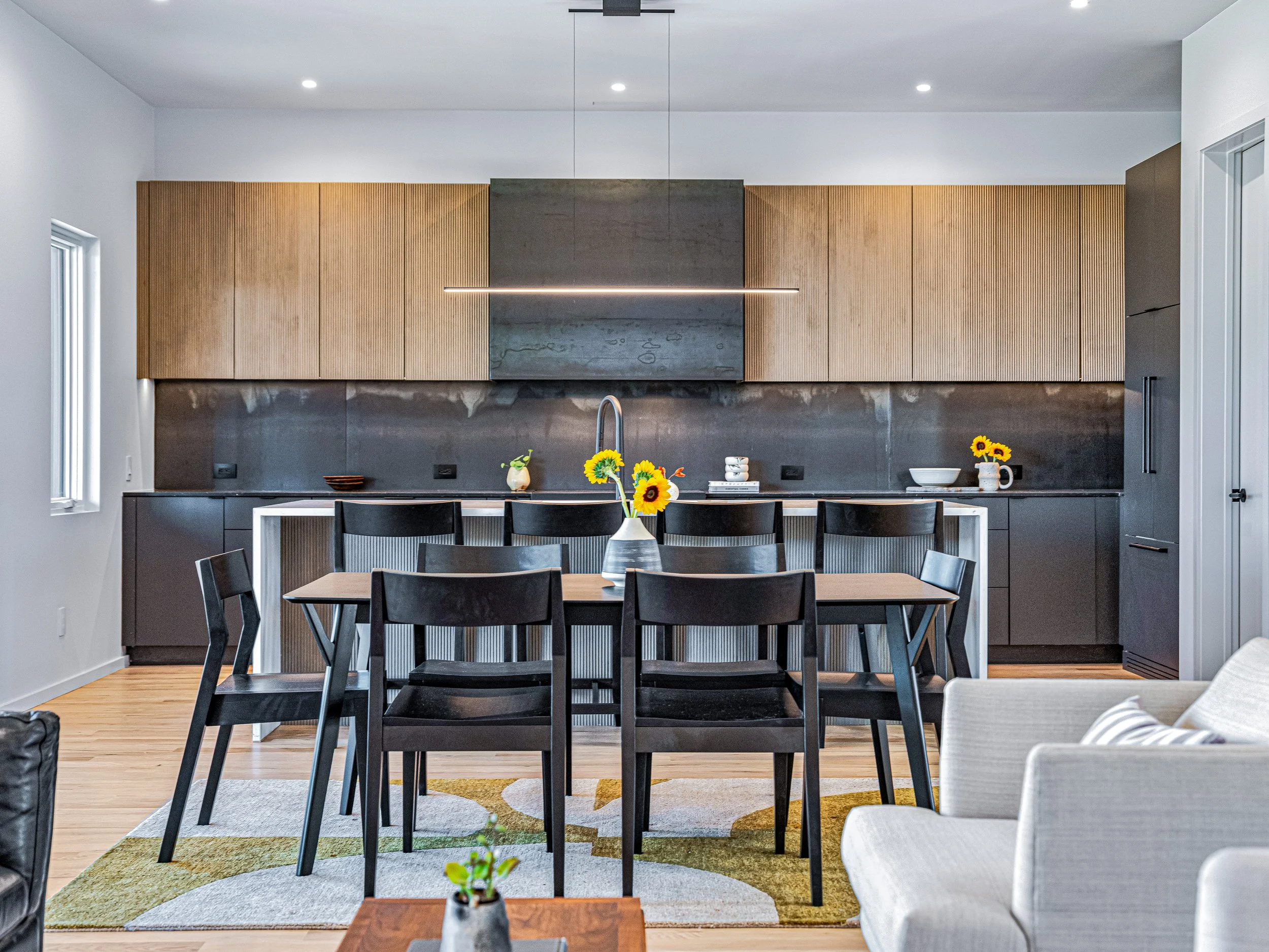 Modern kitchen with black and wood cabinetry, a wooden dining table with six black chairs, and a beige sofa partially visible in the foreground. There are sunflowers in a vase on the table and decor on the kitchen counter. The floor is hardwood, and 