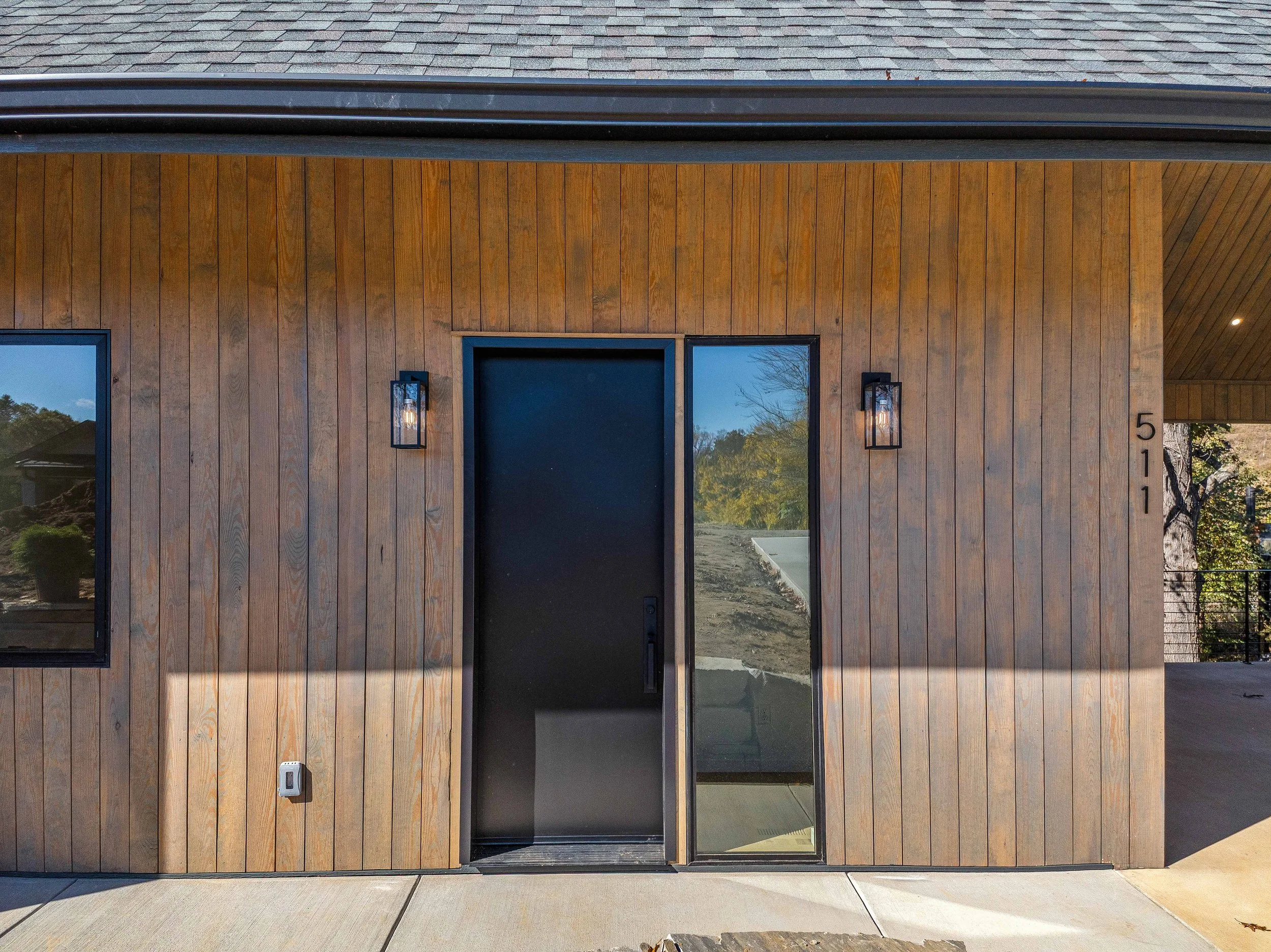 Modern house exterior with wooden siding, black door, side window, wall-mounted lights, house number 511, and surrounding sidewalk and landscape.