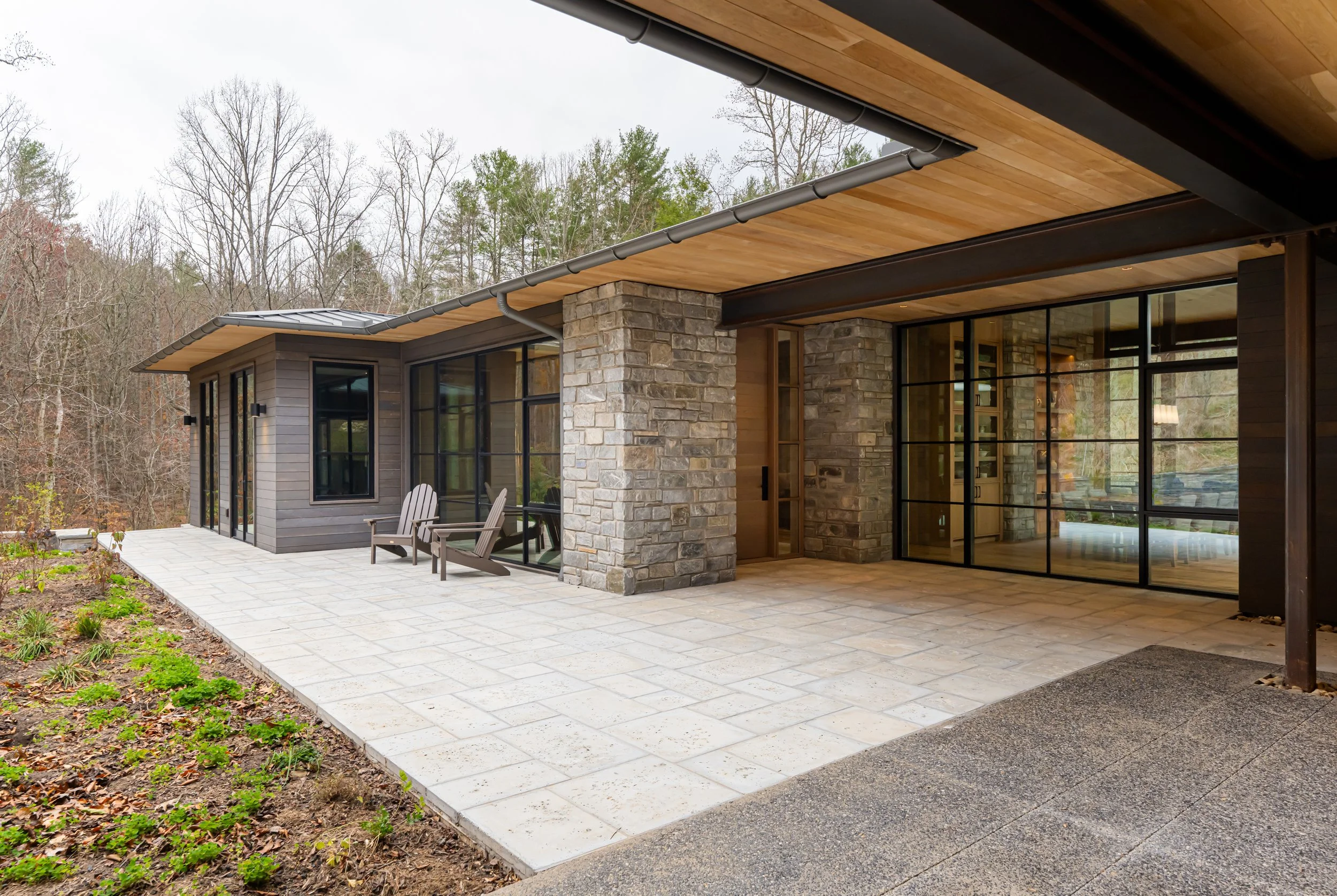 A modern house with a large covered porch, featuring stone and wood siding, black-framed glass doors and windows, and outdoor seating, surrounded by a wooded area with leafless trees.