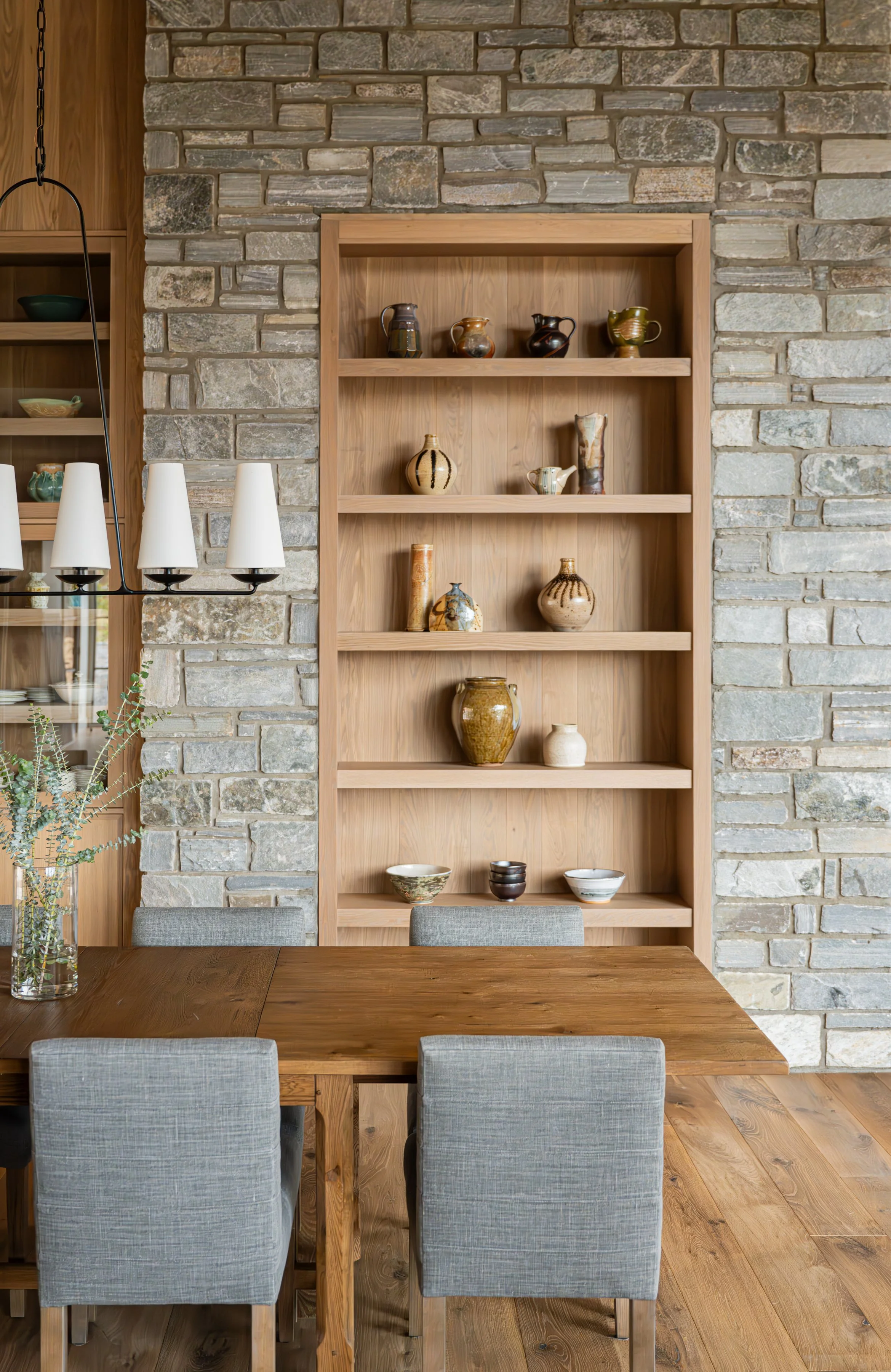 A wooden dining table with four gray upholstered chairs, a glass vase with green branches, and a built-in wooden shelving unit with various ceramic vases and bowls against a stone brick wall.