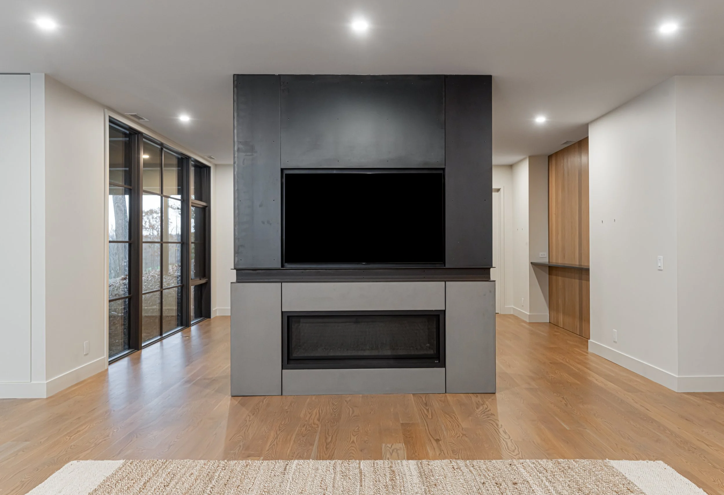Modern living room with a built-in flat-screen TV above a contemporary fireplace, dark-colored wall panels, light wood flooring, large glass windows on the left, and a small wooden accent wall on the right.