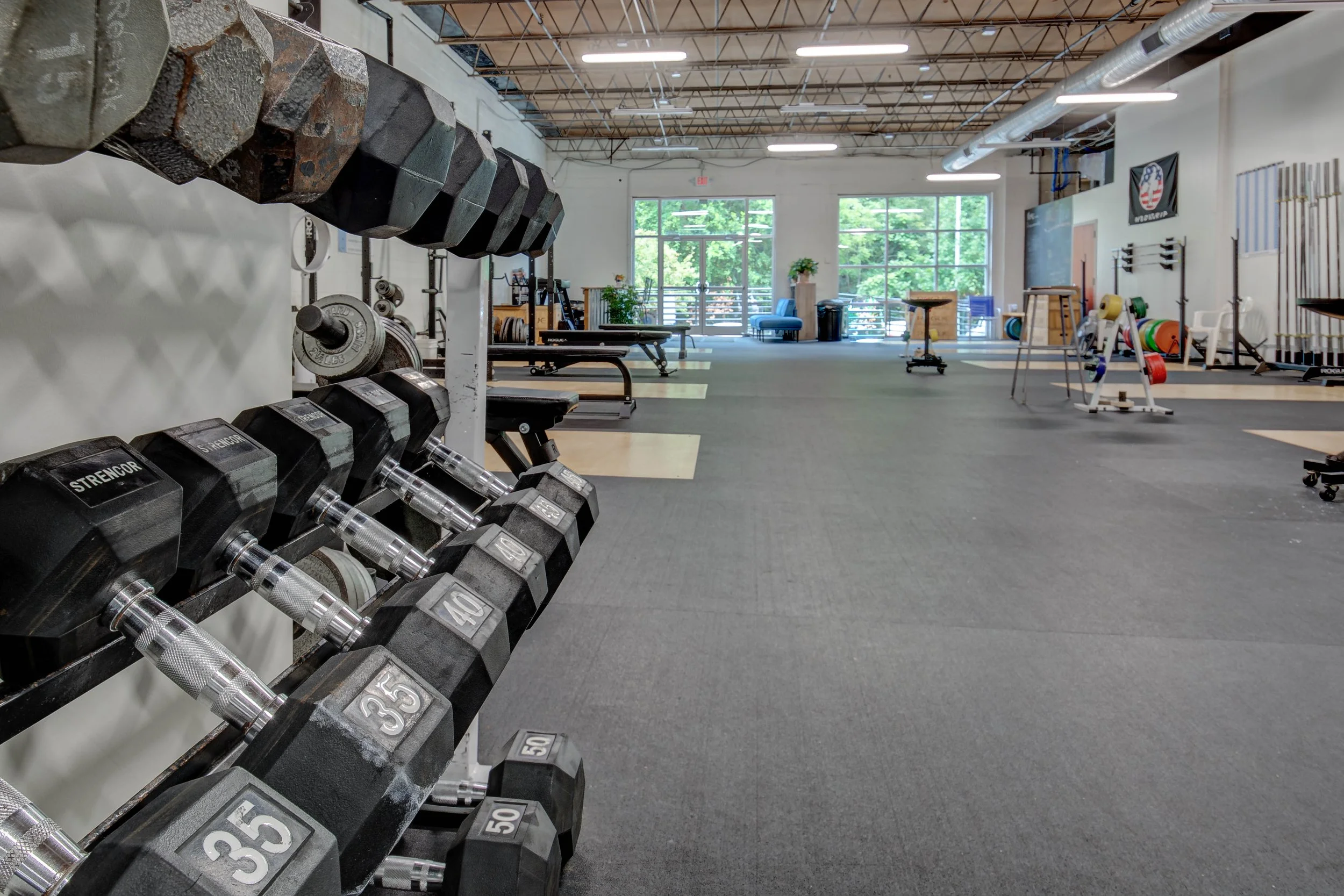 Empty gym with dumbbells on a rack, exercise equipment, benches, and large windows showing green trees outside.