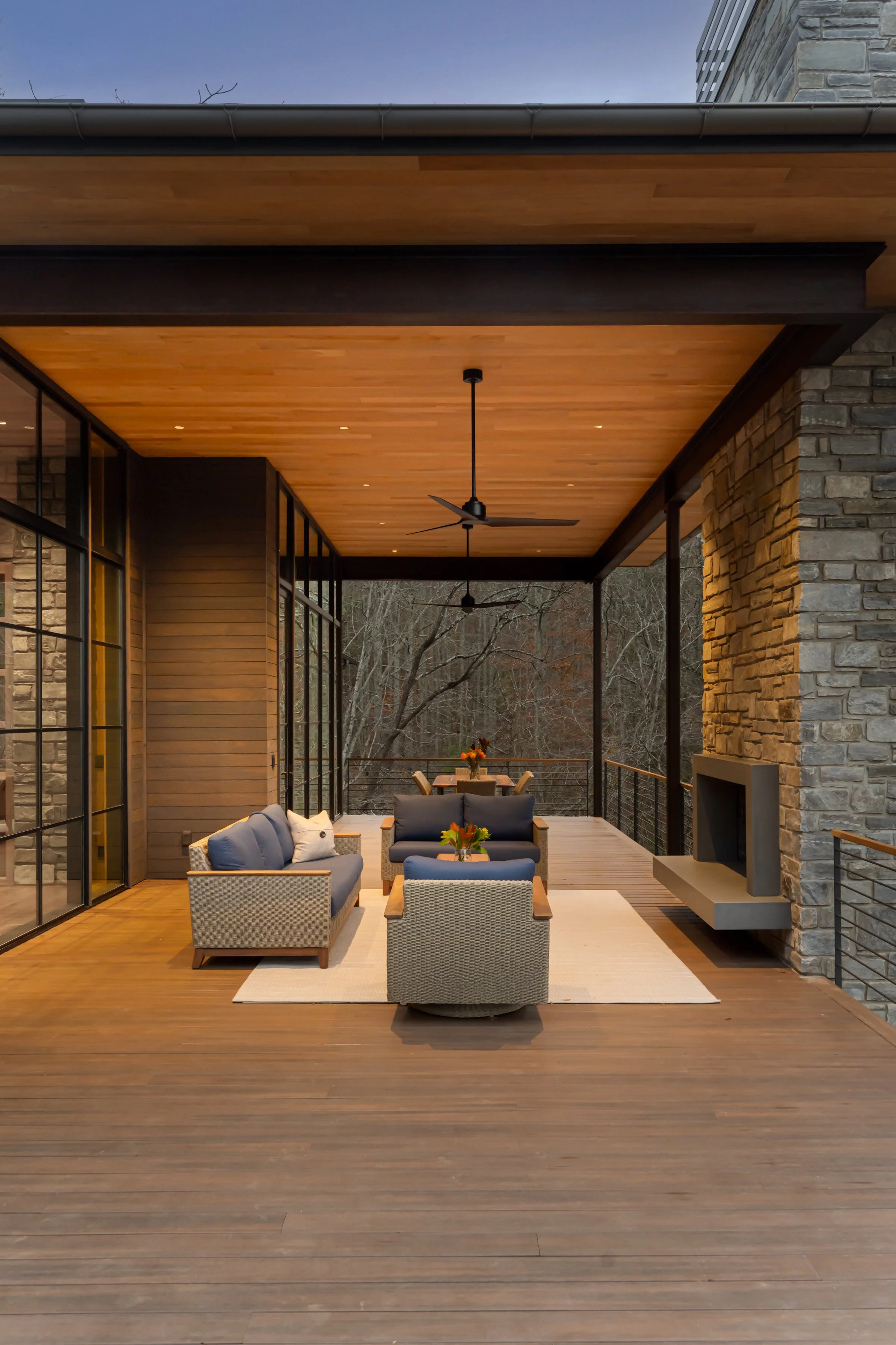Modern outdoor patio with wooden flooring, a ceiling fan, stone fireplace, and outdoor seating including sofas and chairs with cushions.
