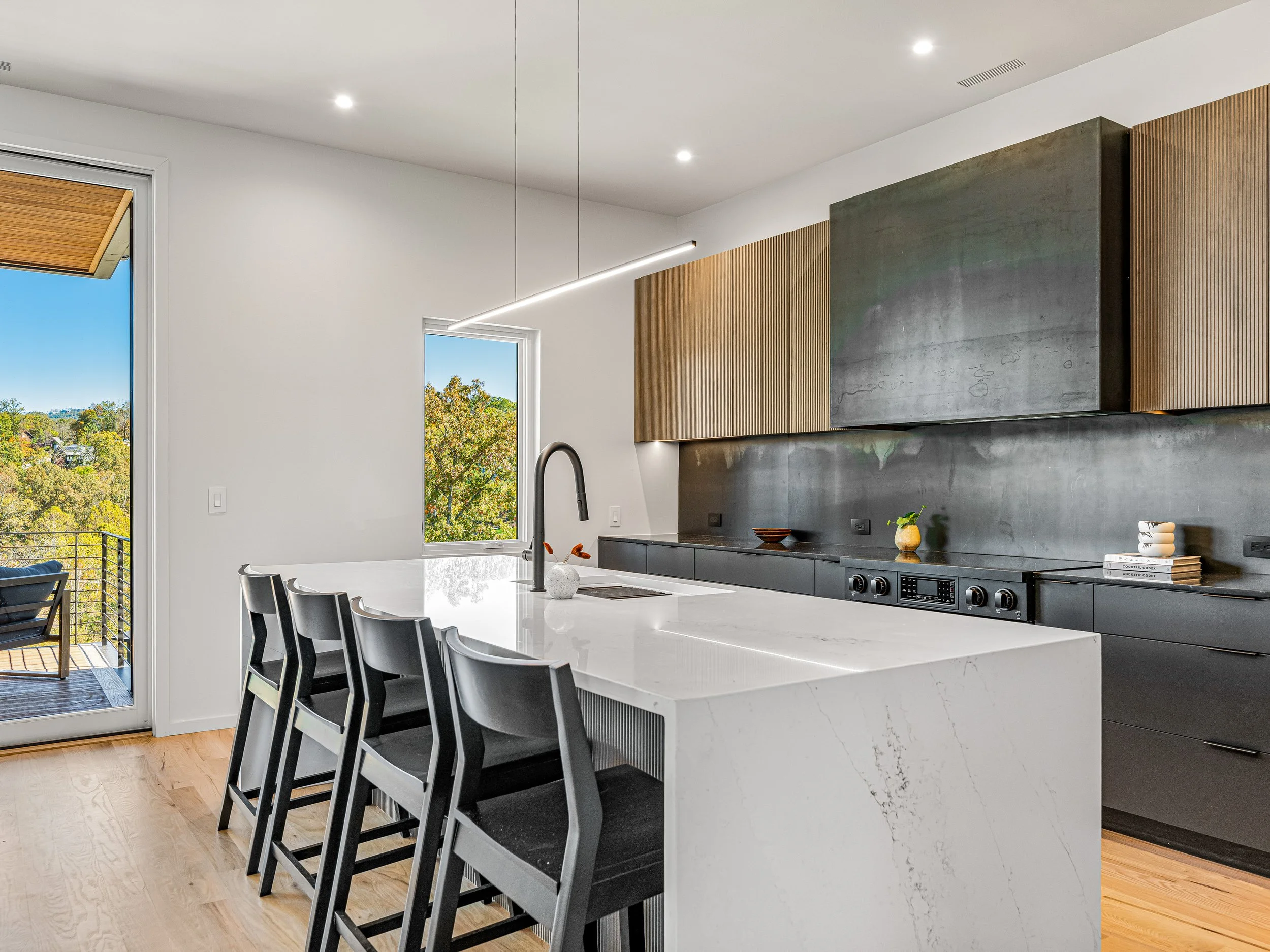 Modern kitchen with white island counter, black chairs, black cabinetry, wooden and black wall cabinets, stainless steel stove, and large windows showing greenery outside