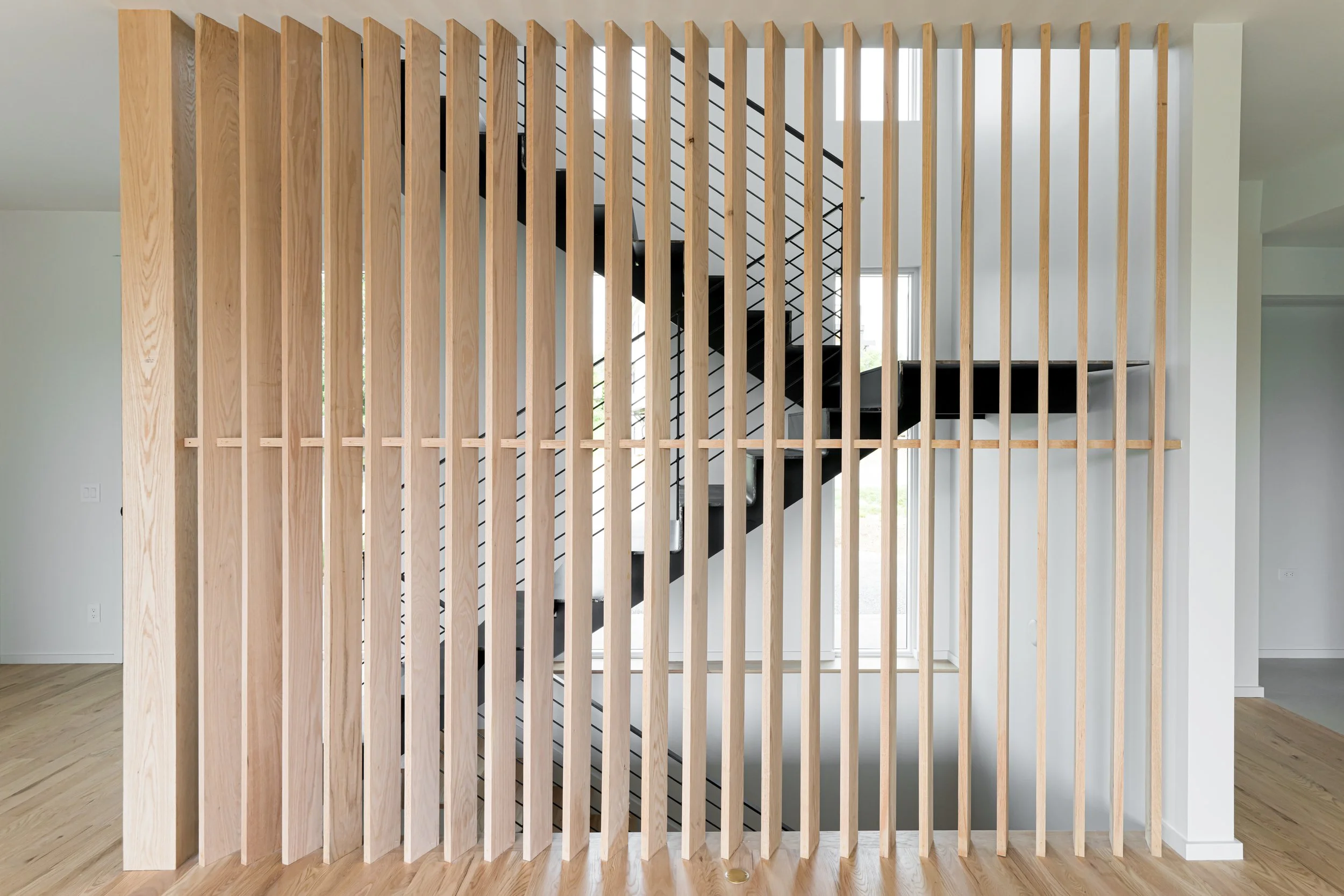 Interior view of a modern home with a staircase seen through vertical wooden slats serving as room divider. The staircase has black steps, white risers, and metal railings, with ample natural light coming through large windows.