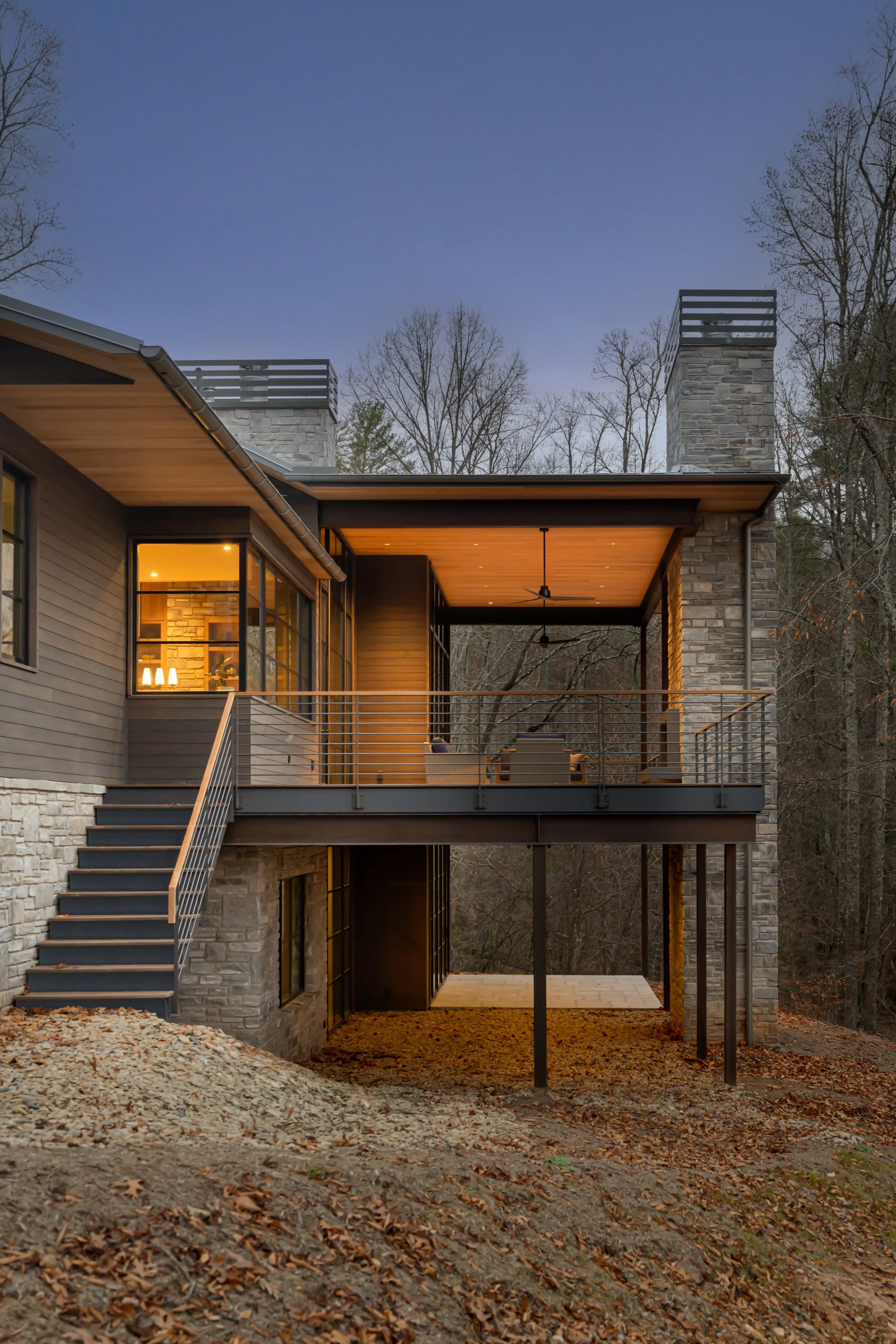 A modern house with a stone and wood exterior, large windows, and a spacious upper balcony illuminated inside, set in a wooded area during dusk.