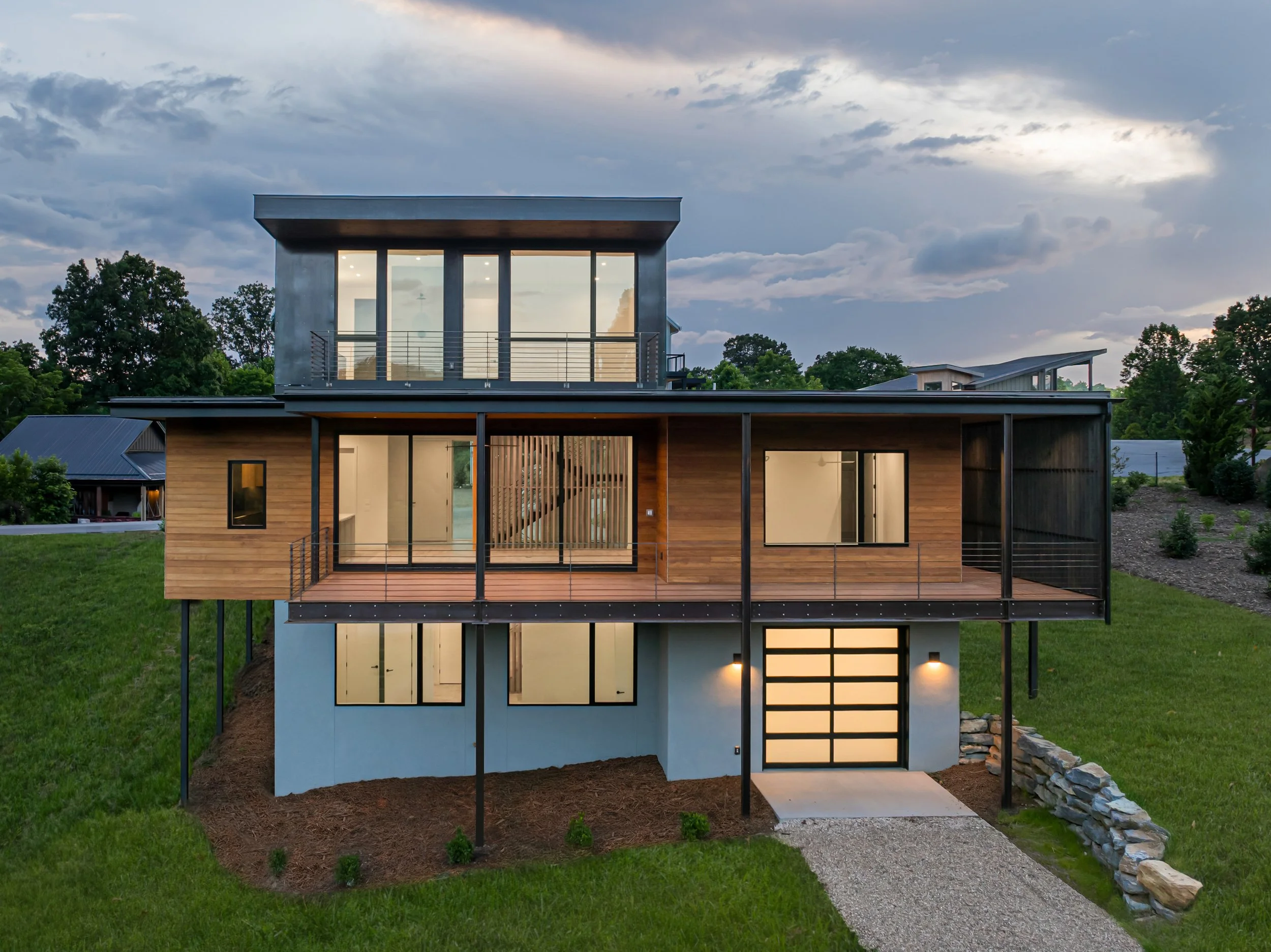 Modern multi-story house with a wooden exterior, large glass windows, and a metal roof, set on a grassy hill at dusk.