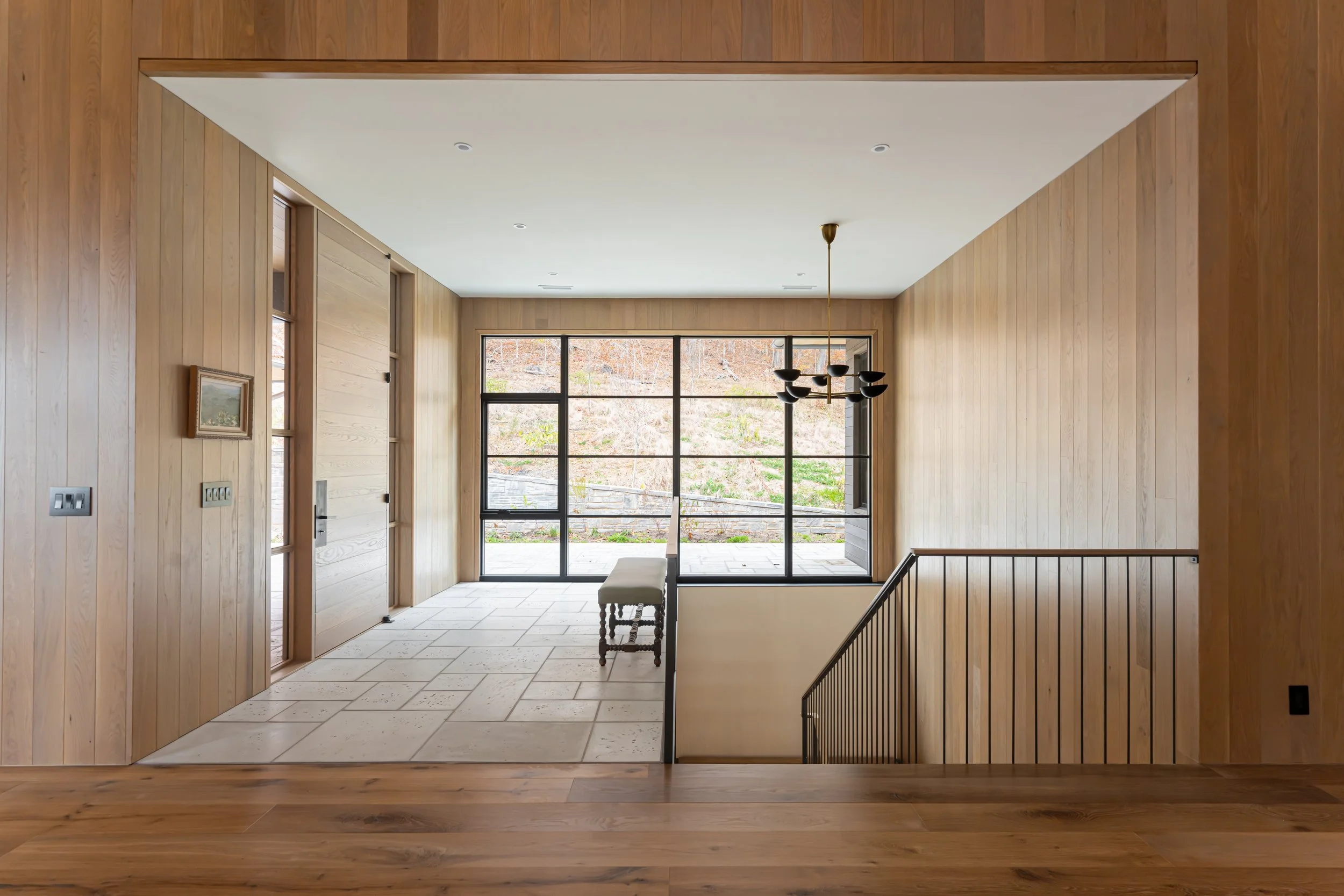Interior view of a modern house, featuring wooden walls, a tiled floor, large glass windows, a bench, a black chandelier, and a staircase railing.