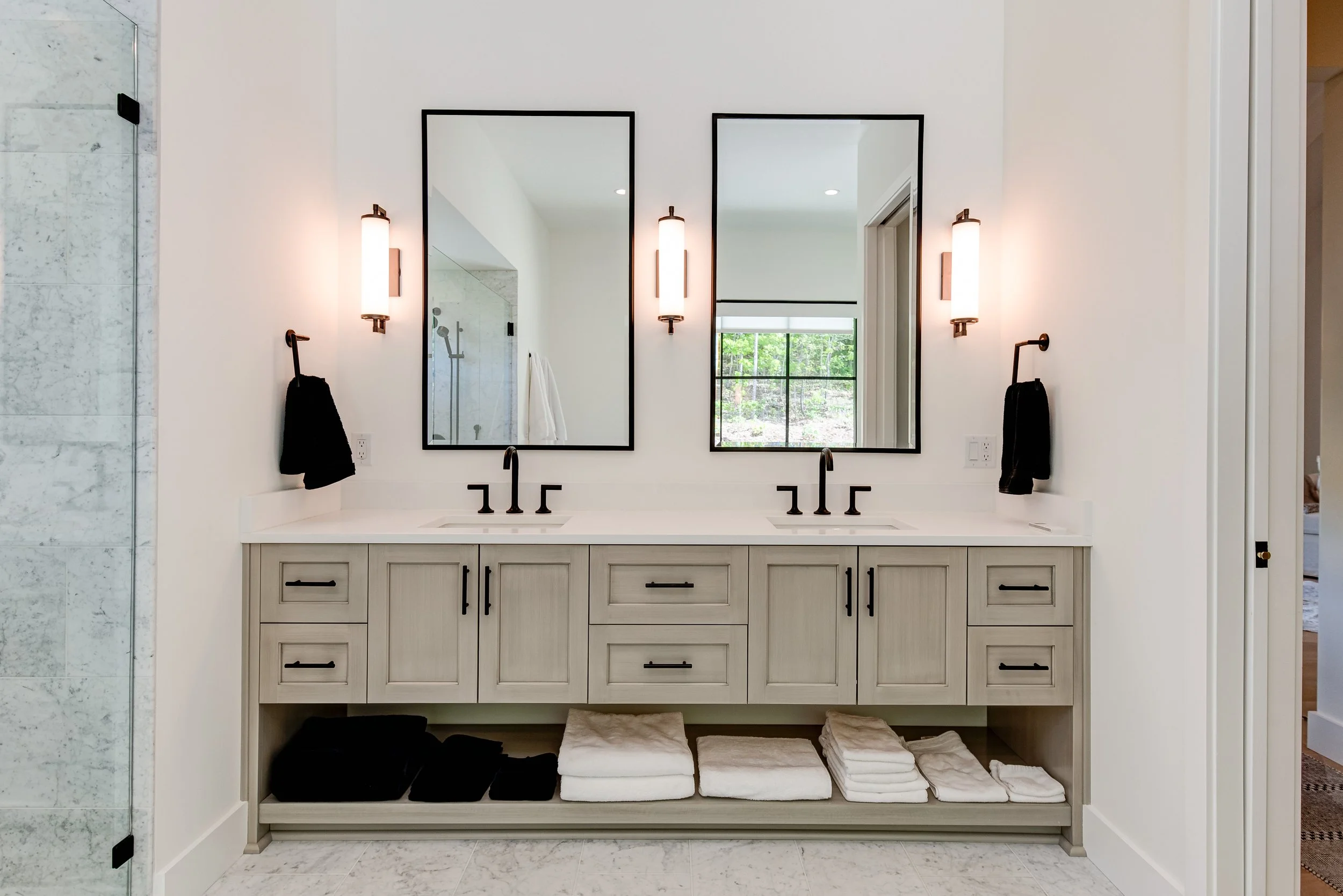 Modern bathroom vanity with two sinks, large mirrors, wall-mounted lights, and black hardware, with towels and black soap dispensers.
