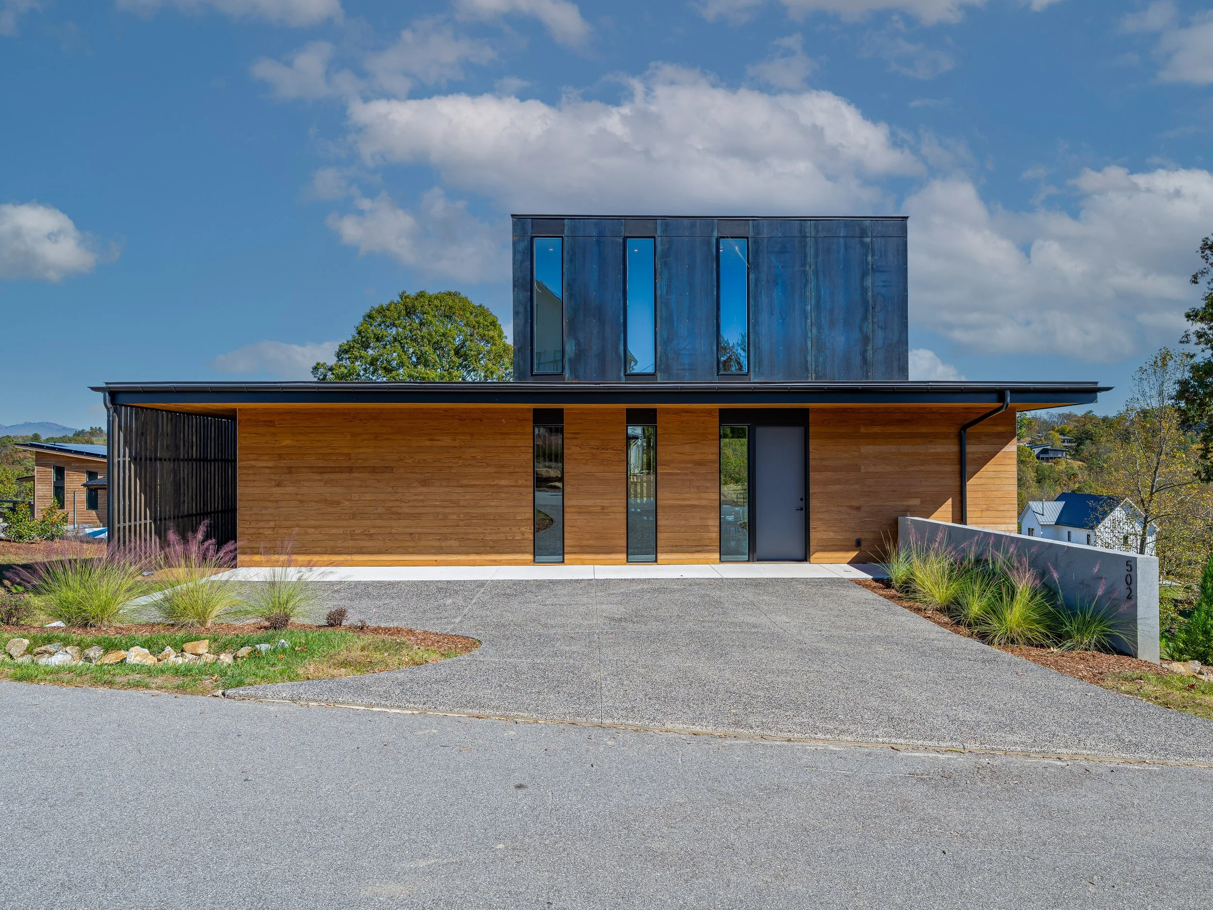 Modern house with wooden and metal exterior, concrete driveway, landscaping with grasses, under a blue sky with clouds.