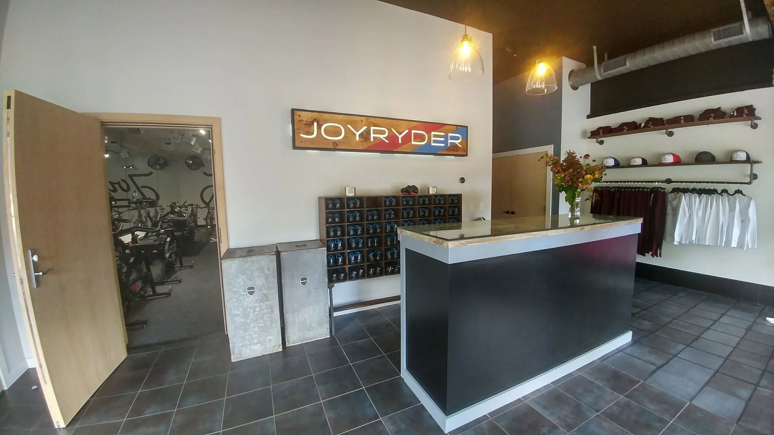 Indoor gym reception area with a black front desk, decorative flowers, and wall-mounted hats and clothing on shelves.