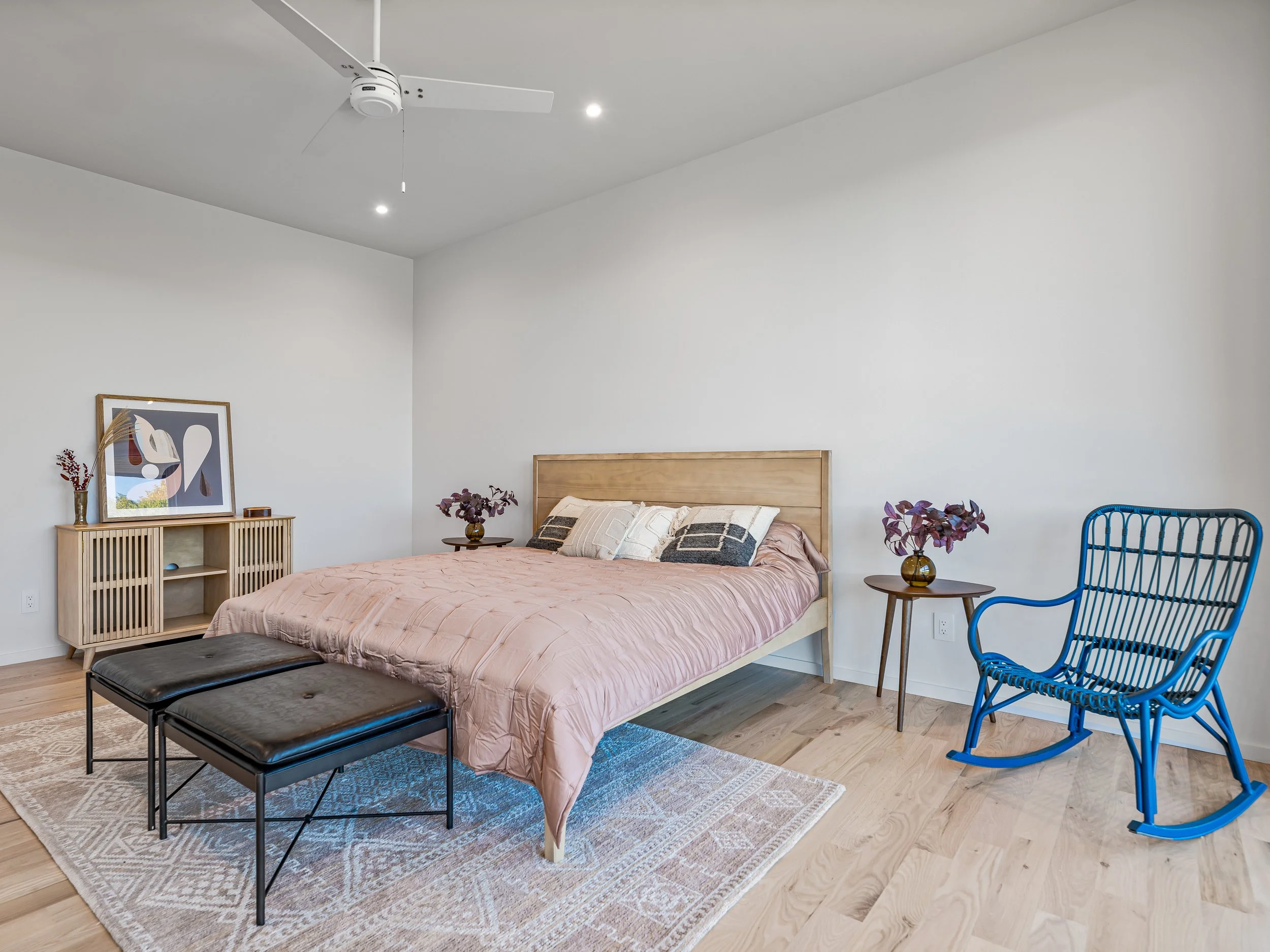 A bedroom with a wooden bed frame, pink bedding, and decorative pillows. Two dark wood nightstands with purple plants. A light wood sideboard with art and a vase. Blue rocking chair, black bench, and a patterned rug on light wood flooring. White wall