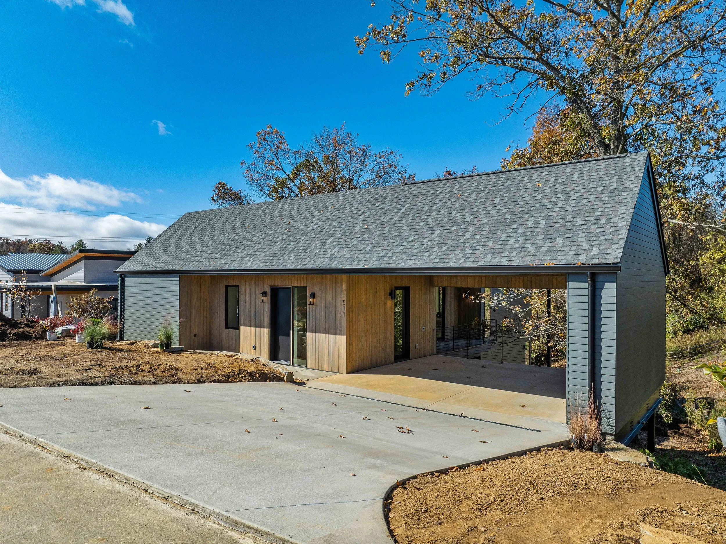 Modern house with a large gabled roof, featuring a combination of gray siding and wooden paneling on the facade, surrounded by a dirt yard and a new concrete driveway, with trees and blue sky in the background.