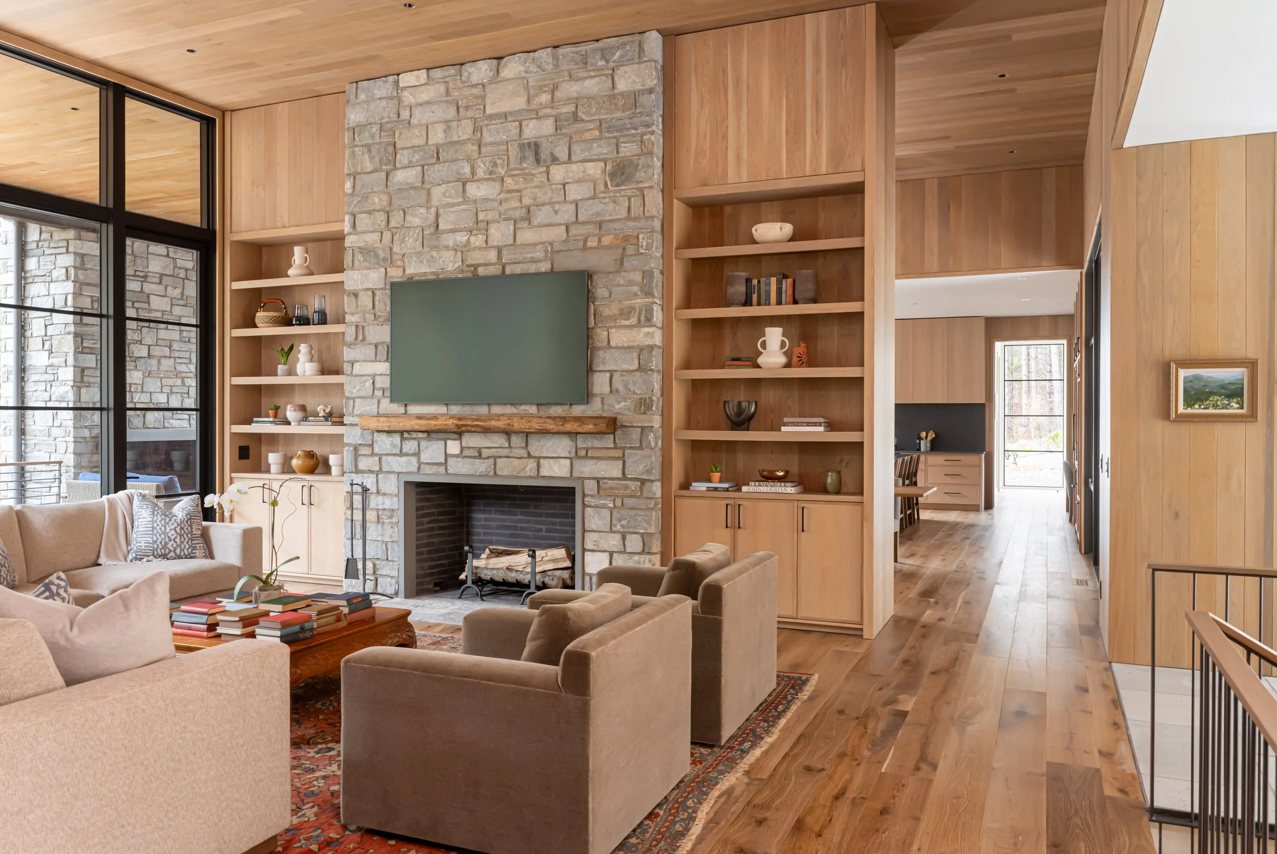 Living room with a stone fireplace, wooden walls, and modern decor, including a beige sofa and armchairs, a wooden coffee table with books, and built-in wooden shelves with decorative items.