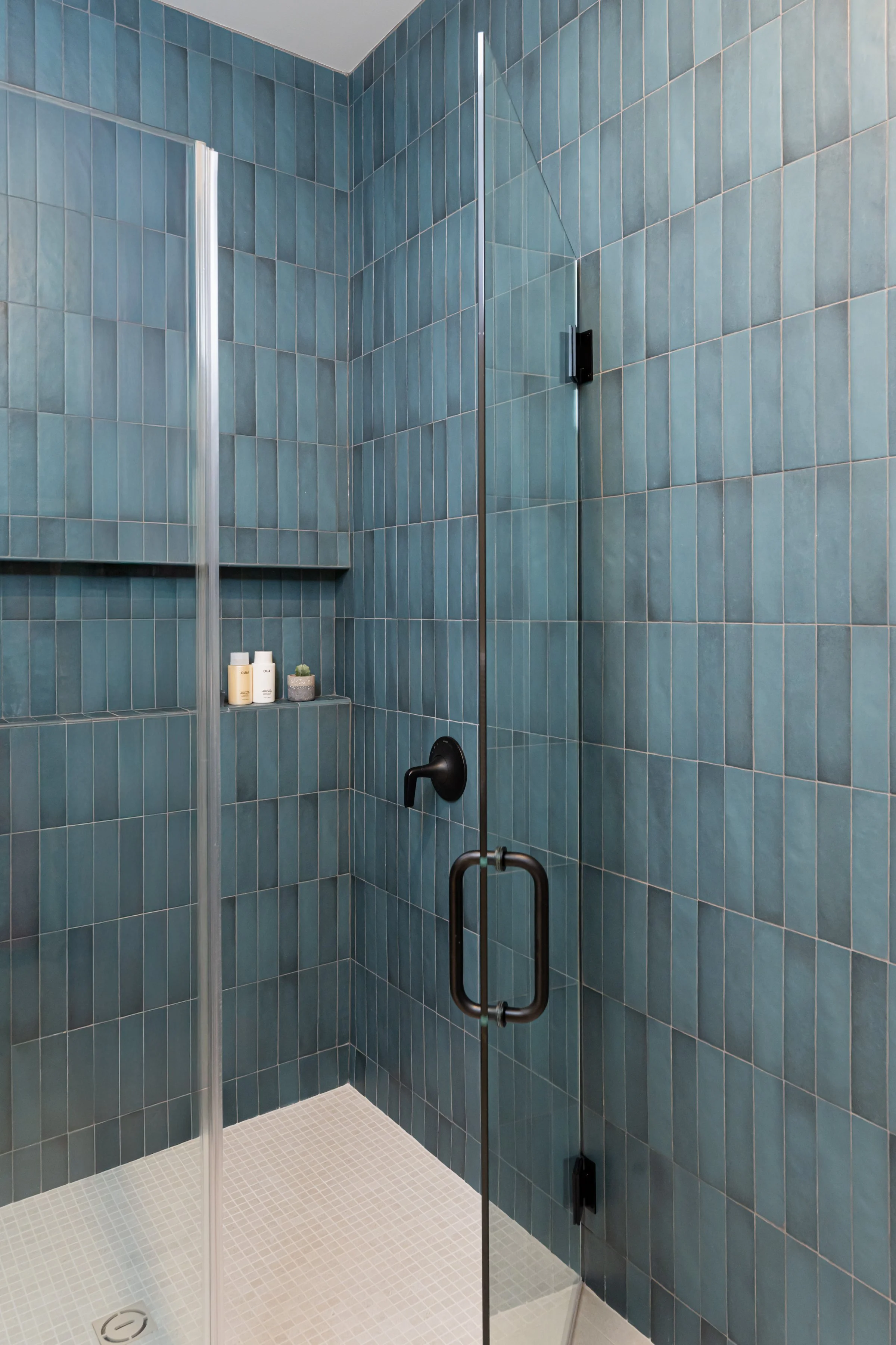 View of a modern shower with teal tile walls, a glass door, a black handle and fixtures, and small shelf holding toiletries.