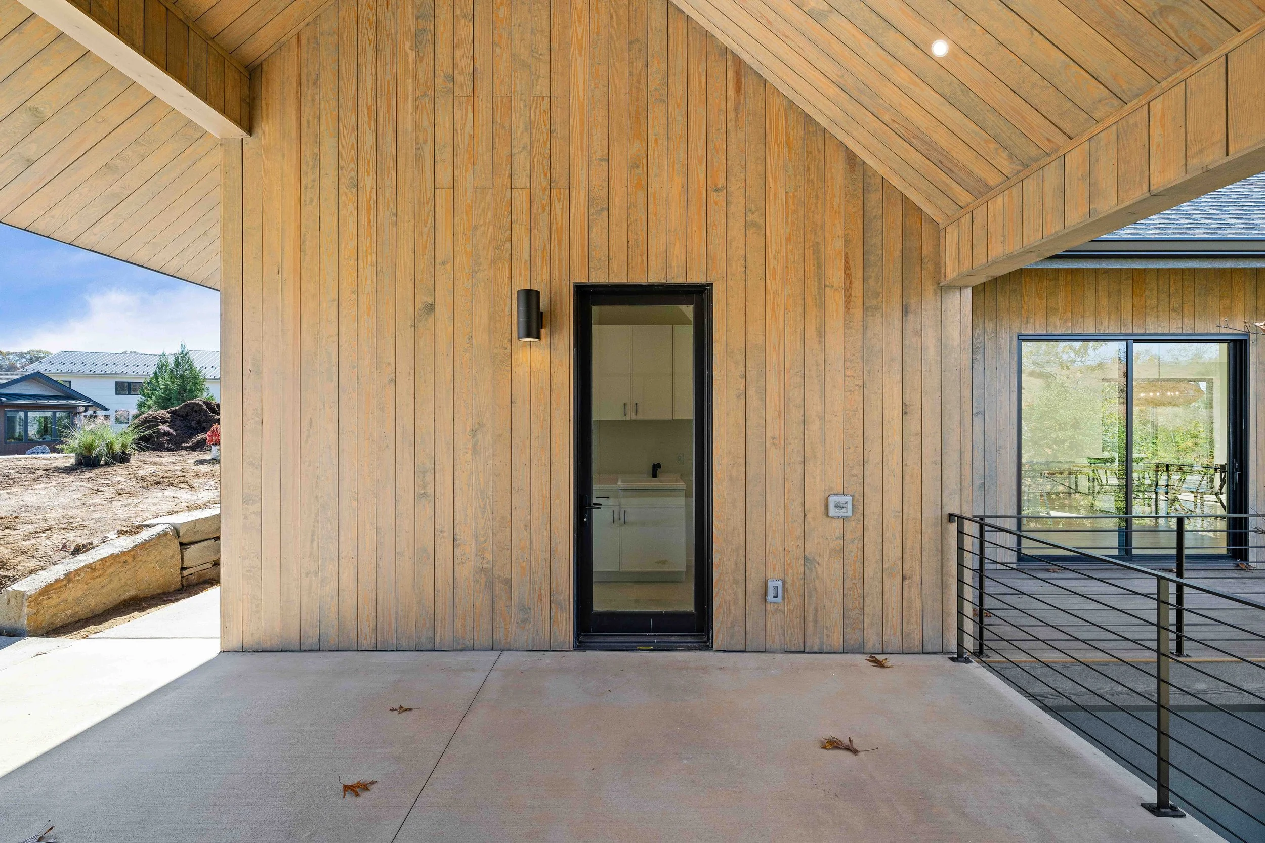 Exterior view of a house with wooden siding, a black framed glass door leading to a laundry room, a black wall-mounted light fixture, and a sliding glass door with a metal railing on a covered porch.