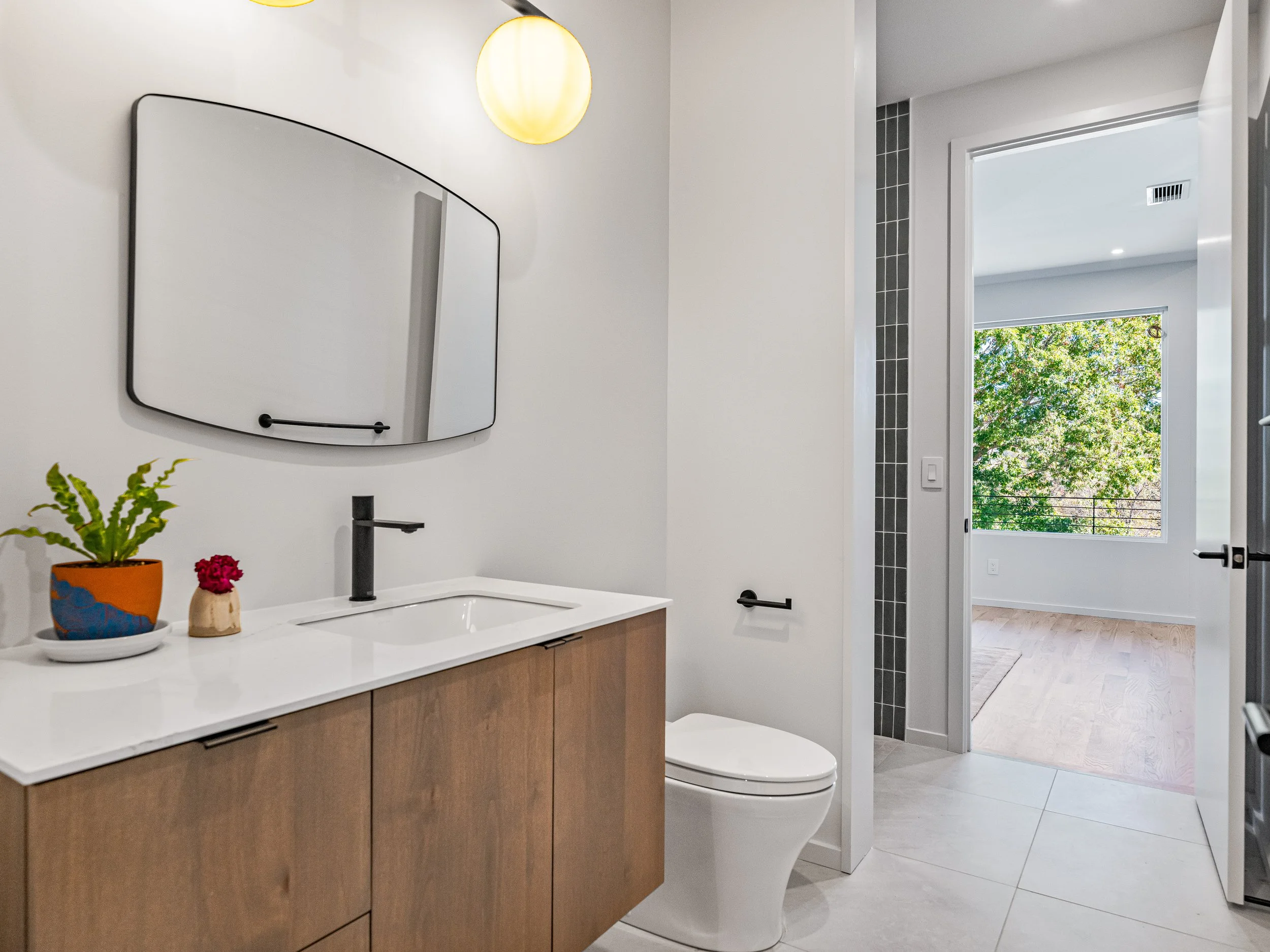 Modern bathroom with a wooden vanity, white countertop, black faucet, oval mirror, wall-mounted light fixture, potted plant, small decorative item, toilet, and view of a bright room with large window and trees outside.