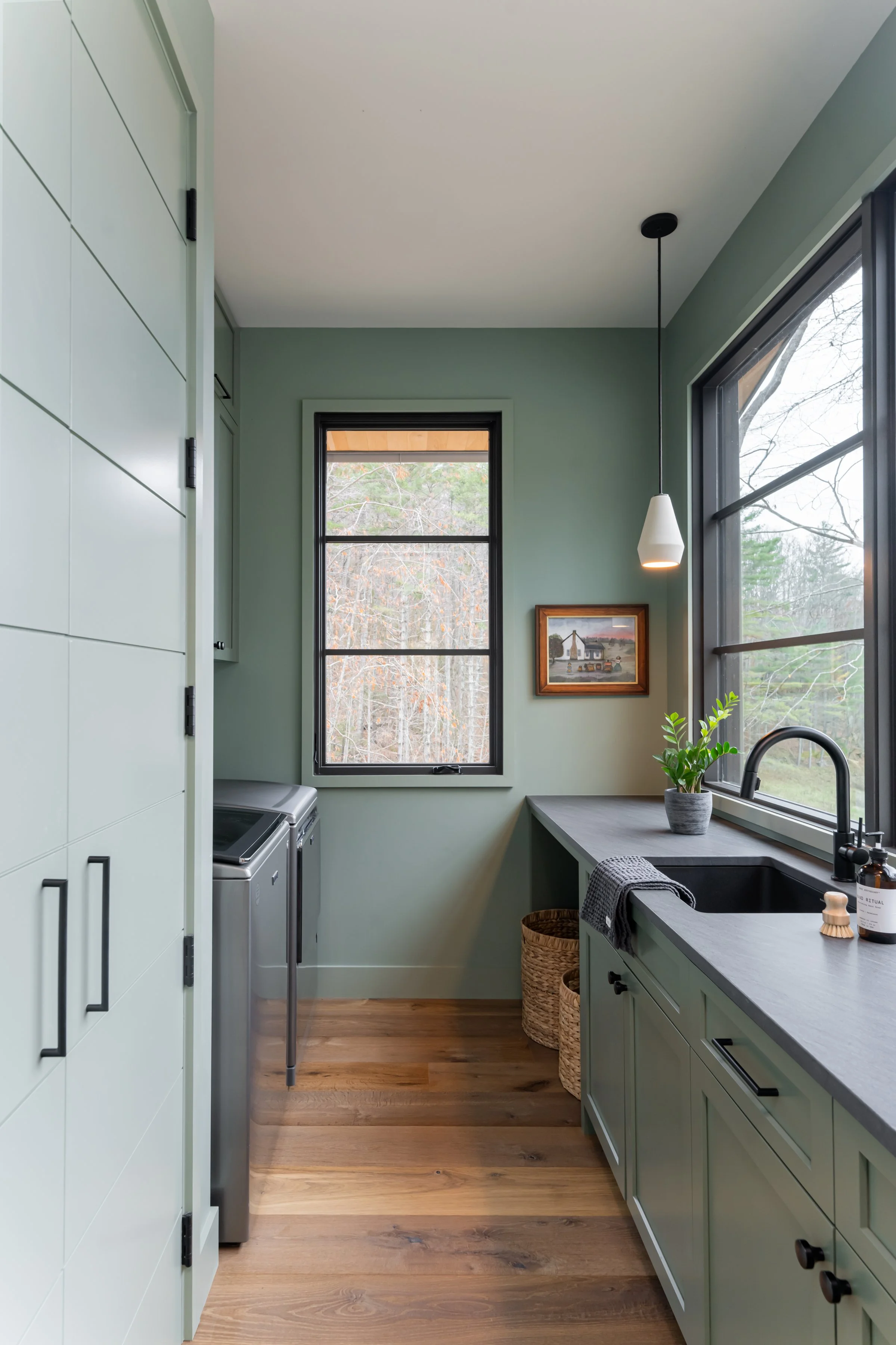 A laundry room with a green cabinet, black countertops, a window, a washing machine, a plant, and wooden baskets.