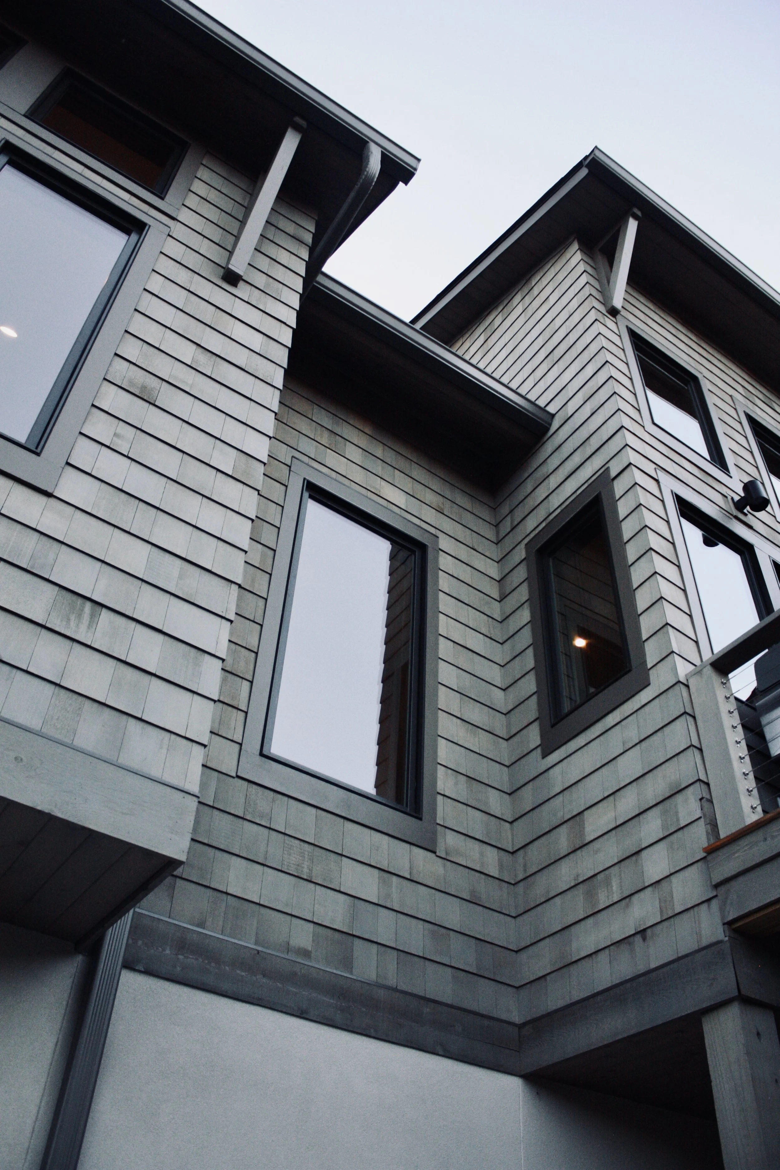 Close-up of modern house exterior with gray siding, large windows, and overhanging roof.
