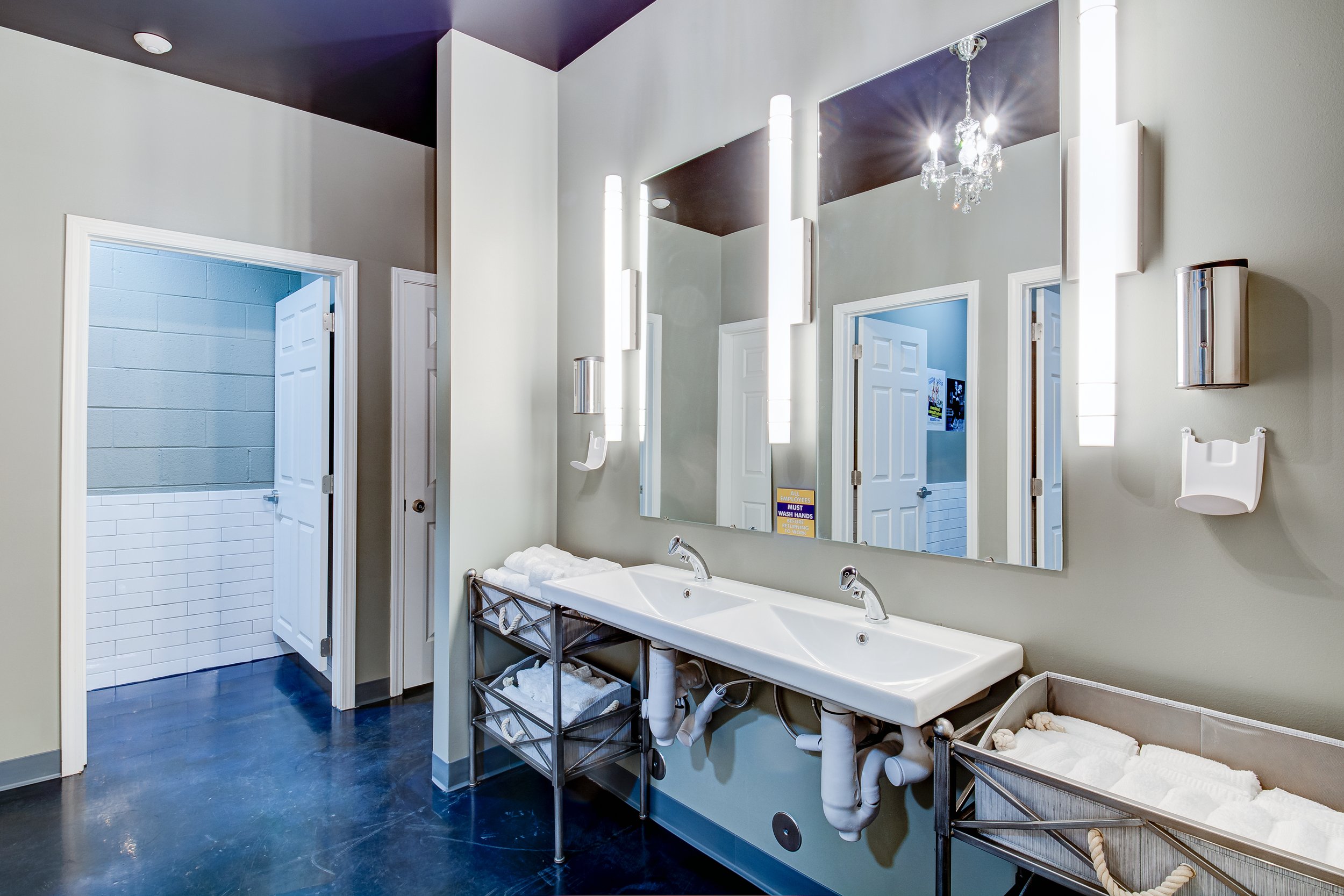A modern bathroom with dual sinks, large mirror, and a chandelier. The bathroom has a walk-in shower with a blue and white tile design. There are towels on a metal shelf next to the sinks and a soap dispenser on the wall.
