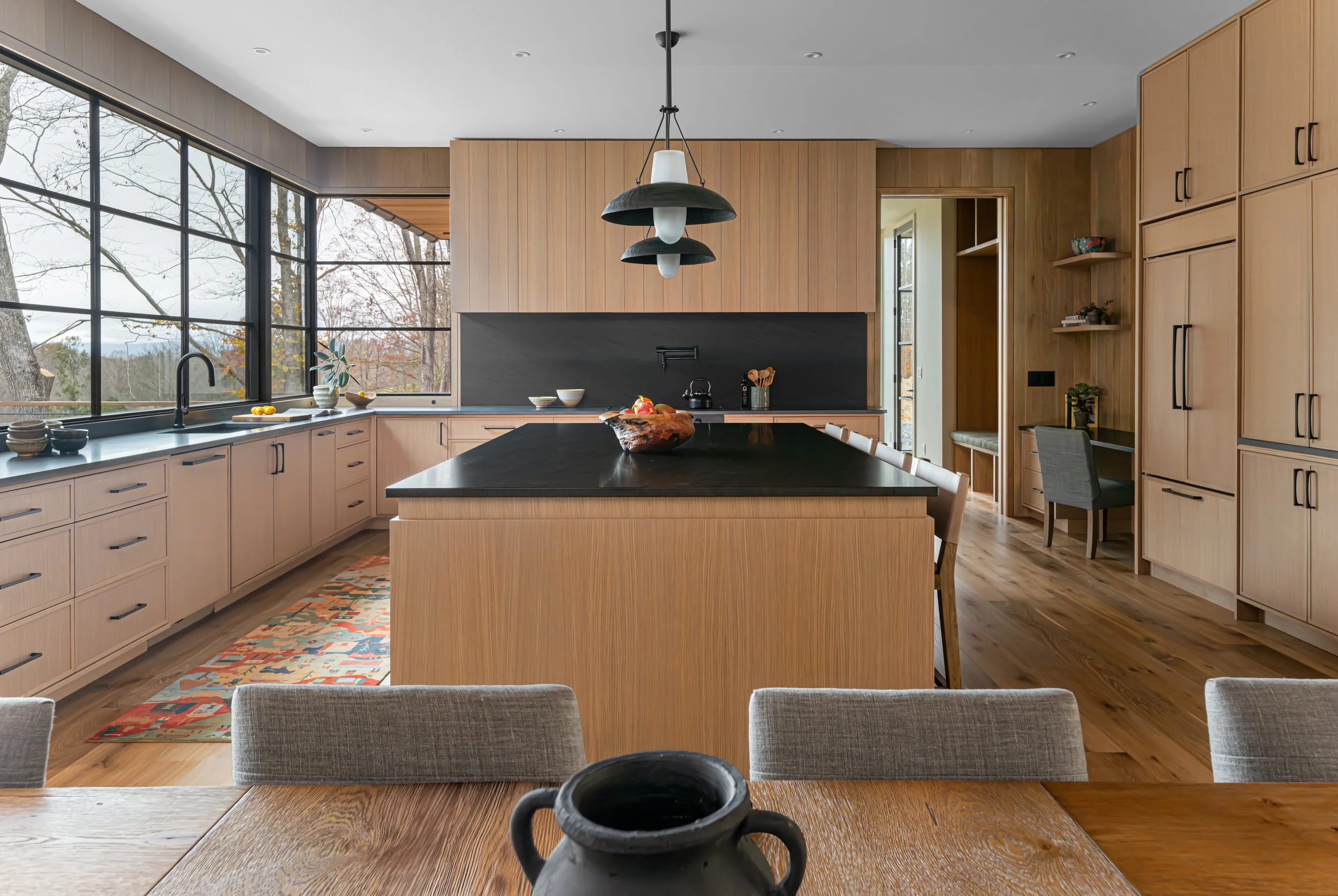 Modern kitchen with light wood cabinets, black countertops, a large island, a black hanging light fixture, large windows showing trees outside, and a dining table with chairs in the foreground.